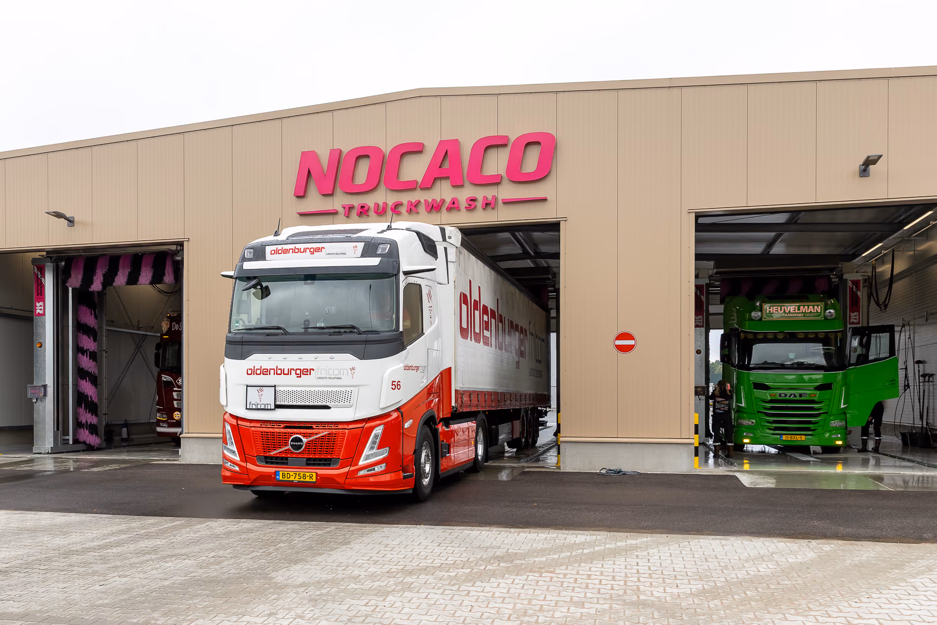 Een rode en witte vrachtwagen met het logo 'Oldenburger' rijdt uit een wasstraat van Nocaco Truckwash, naast een groene vrachtwagen binnen de wasstraat.