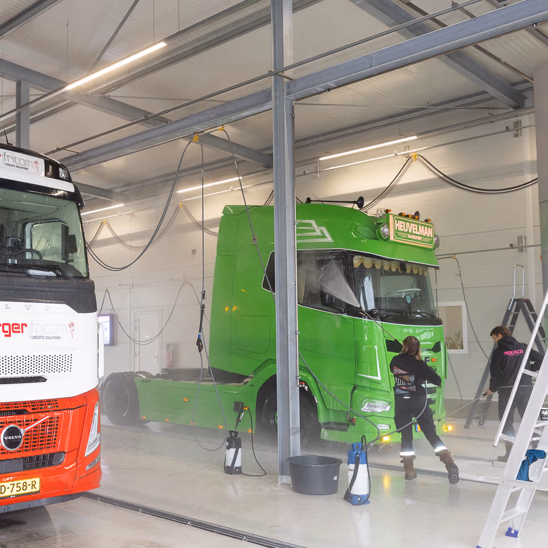 Twee grote vrachtwagens staan in een lichte werkplaats: een witte en rode Volvo-vrachtwagen aan de linkerkant en een groene vrachtwagen met HEUVELMAN-bord aan de voorkant. Twee mensen in donkere werkkleding zijn bezig bij de groene vrachtwagen, één dichtbij en één bij een metalen trapladder rechts.