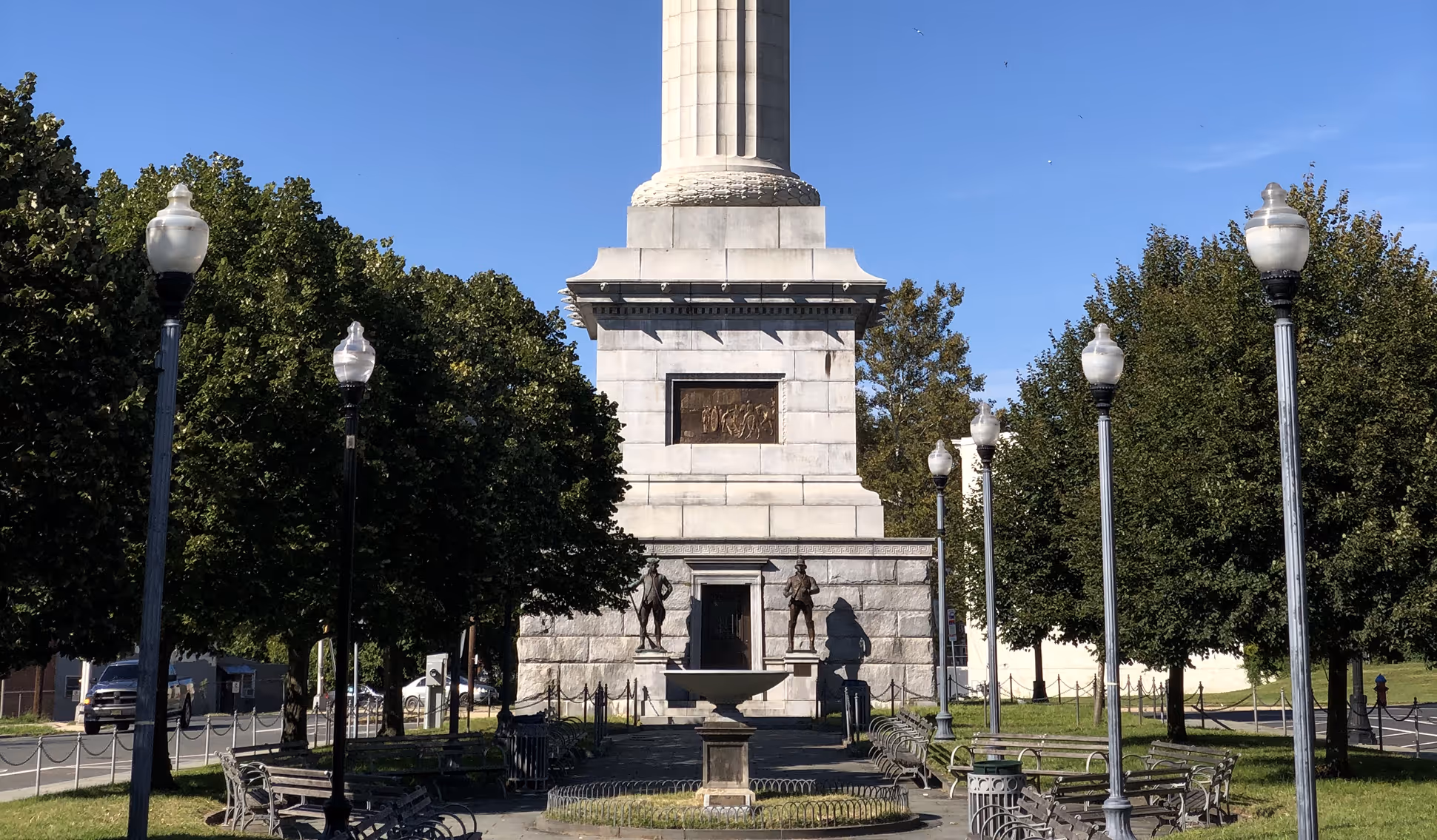 North Trenton Battle monument with two bronze statues, central fountain, benches, and lampposts in a tree-lined park under a clear blue sky.