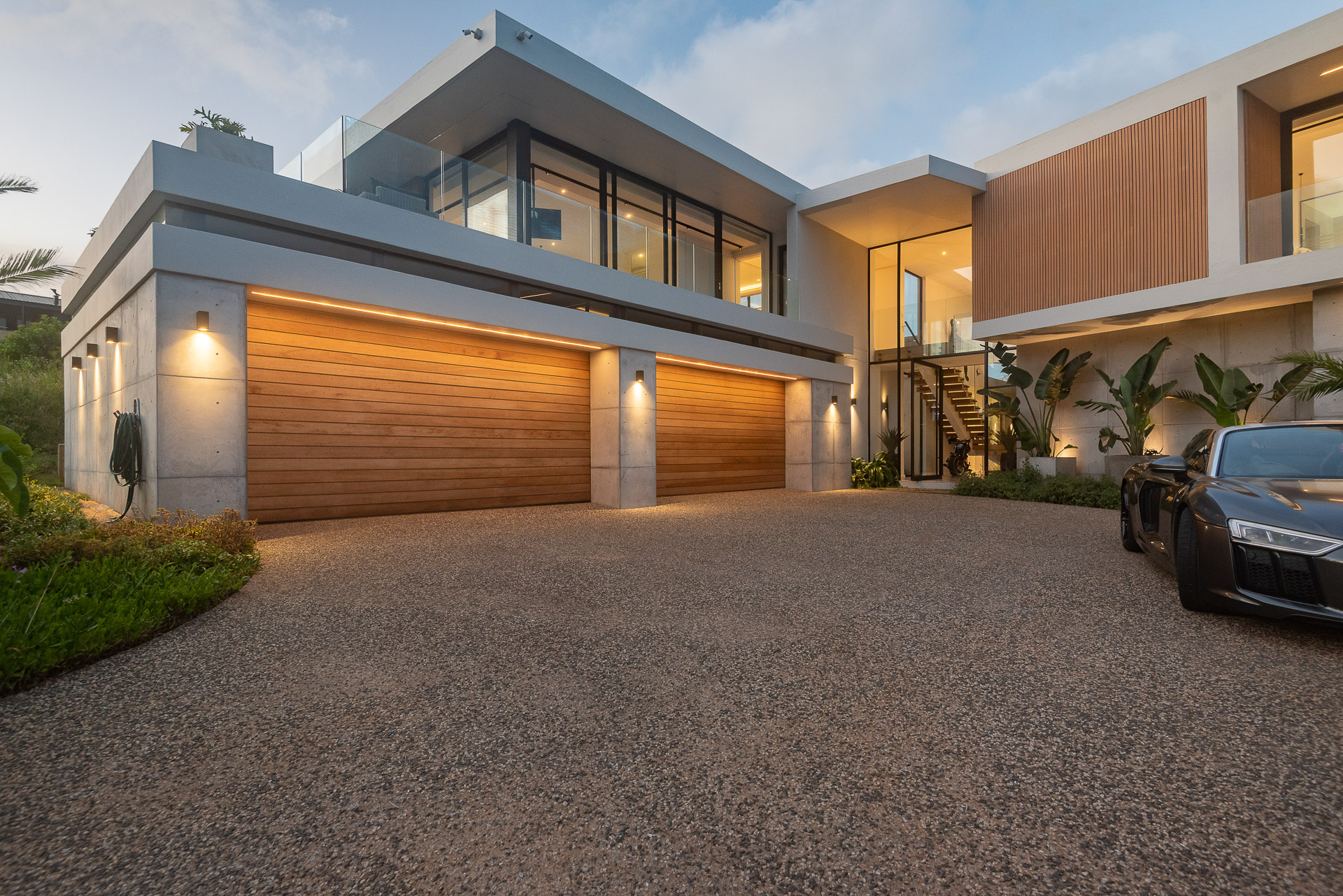 Exterior arrival view of House Django, a bespoke contemporary home designed by Gerhard Architect in Zululami Luxury Coastal Estate on KwaZulu-Natal's North Coast. The image captures the home's sculptural street façade, featuring floating roof planes, warm timber garage doors, exposed concrete columns, expansive corner glazing, a double-height glazed entrance revealing the floating staircase, integrated architectural lighting, tropical gardens and a textured aggregate driveway that creates a welcoming arrival experience.