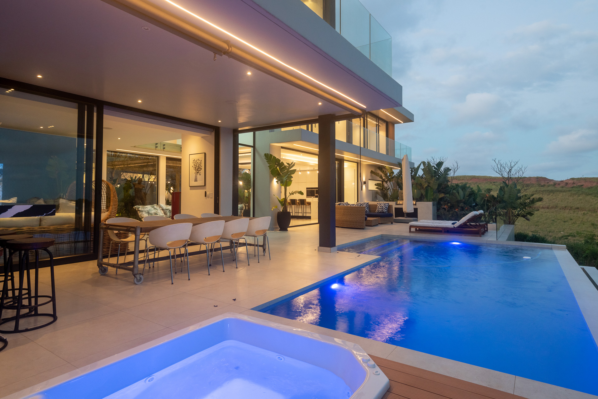Twilight view of the outdoor entertainment area at House Django, a bespoke contemporary home designed by Gerhard Architect in Zululami Luxury Coastal Estate. The photograph features a covered alfresco dining terrace, illuminated infinity pool and spa, floor-to-ceiling sliding glass doors, integrated architectural lighting and open-plan living spaces that extend effortlessly into the surrounding coastal landscape, embodying the home's modern indoor-outdoor lifestyle.