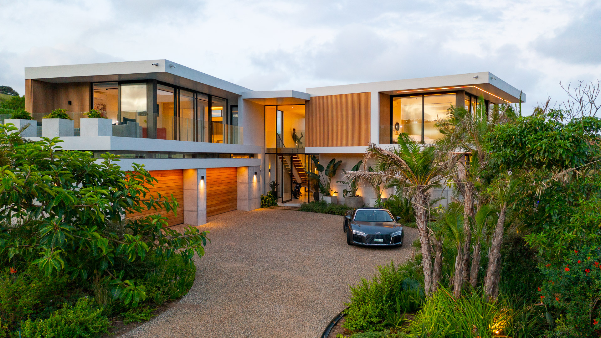 Elevated exterior view of House Django, a contemporary luxury home designed by Gerhard Architect in Zululami Luxury Coastal Estate on KwaZulu-Natal's North Coast. The photograph captures the residence nestled within mature indigenous landscaping, featuring cantilevered roof planes, warm timber cladding, floor-to-ceiling glass, exposed concrete elements, a central glazed entrance with floating staircase and a landscaped arrival courtyard that blends contemporary architecture with the surrounding coastal environment.