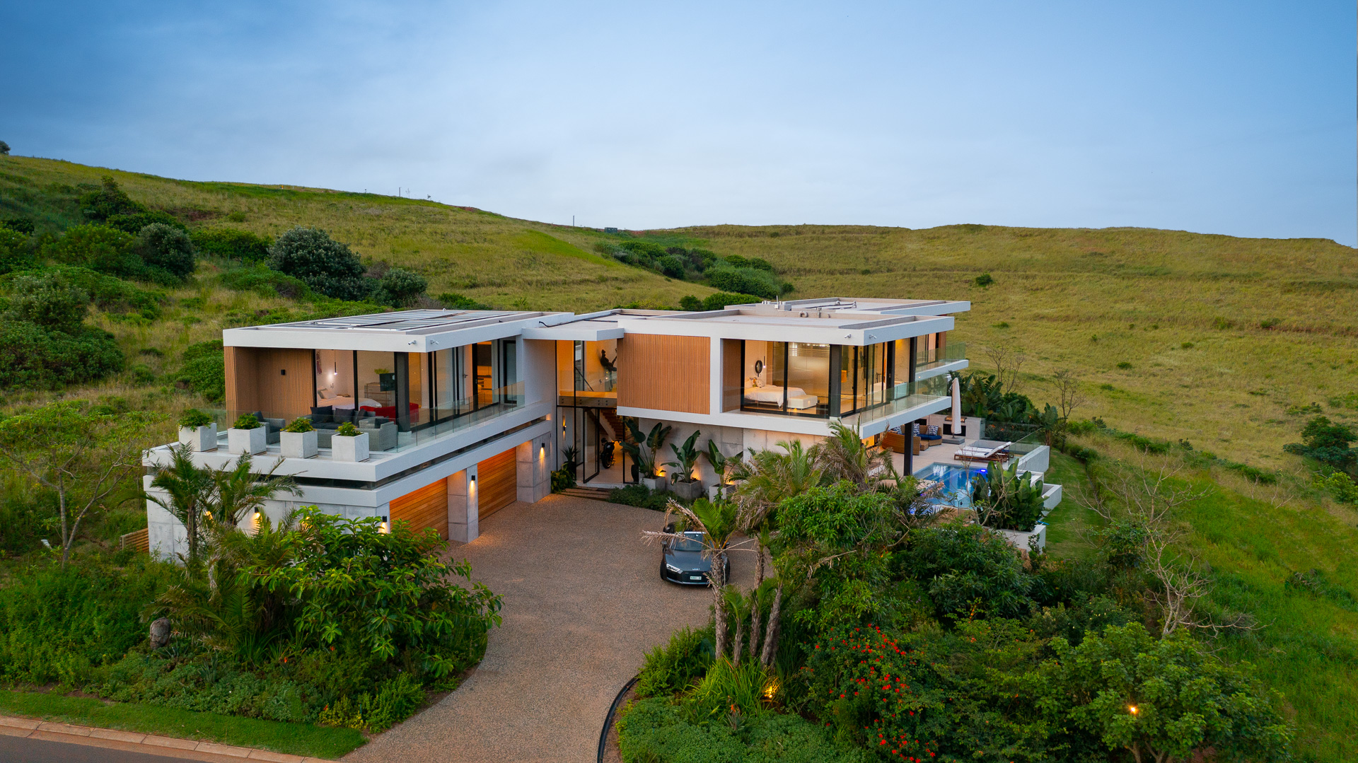 Elevated aerial photograph of House Django, a contemporary luxury home designed by Gerhard Architect in Zululami Luxury Coastal Estate. The image captures the residence nestled within indigenous landscaping, featuring cantilevered roof forms, wraparound glazed balconies, a central double-volume entrance, infinity pool, tropical gardens and a curved aggregate driveway, demonstrating the home's sensitive response to its elevated coastal setting and uninterrupted greenbelt views.