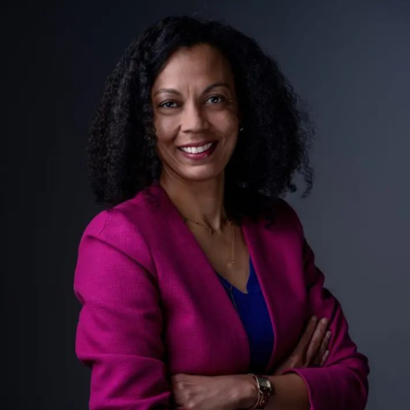 Smiling woman with curly black hair wearing a magenta blazer over a blue top, posed with arms crossed against a dark gray background.