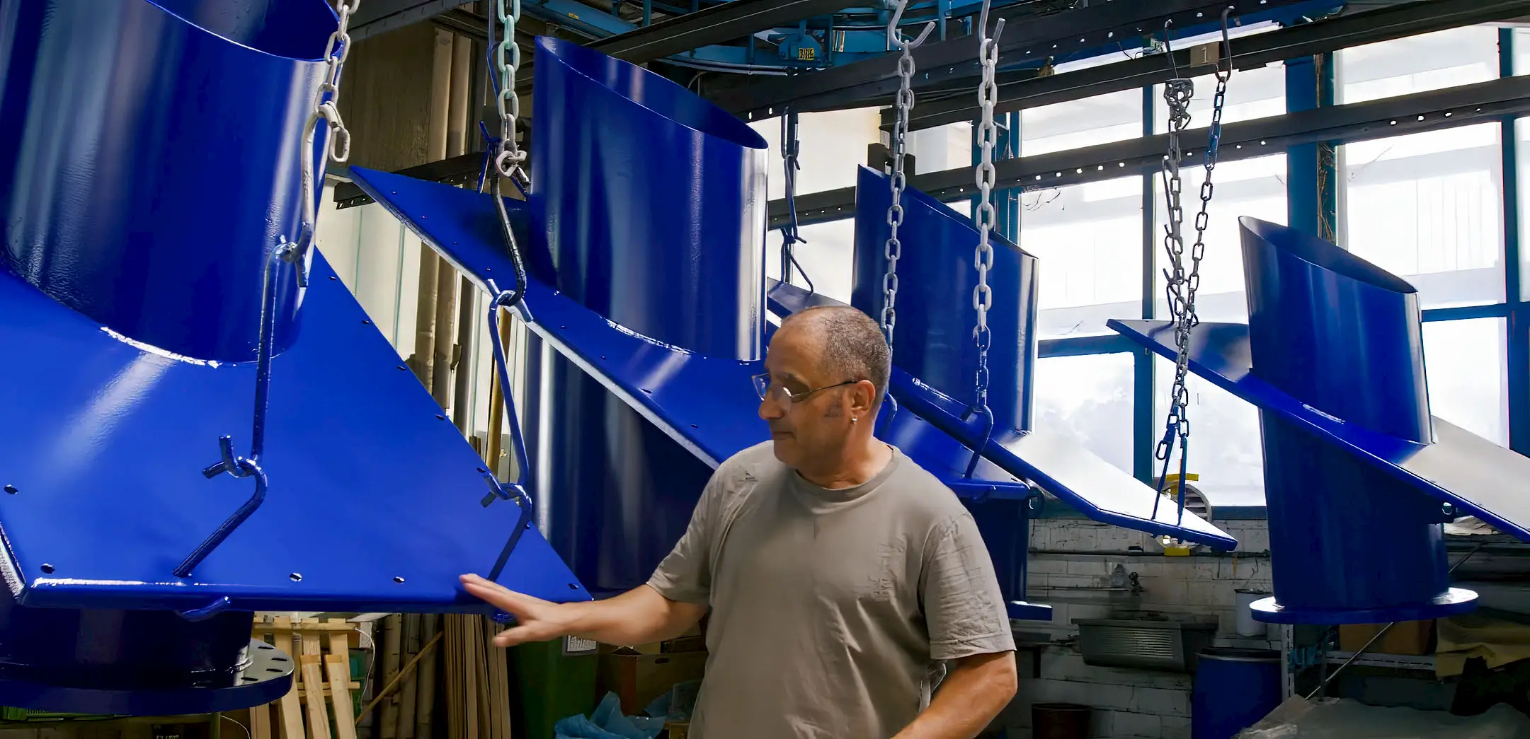 Mitarbeiter kontrolliert in einer Produktionshalle frisch blau beschichtete Metallteile, die an Ketten aufgehängt sind. Die glänzenden Oberflächen zeigen eine hochwertige und gleichmässige Pulverbeschichtung.