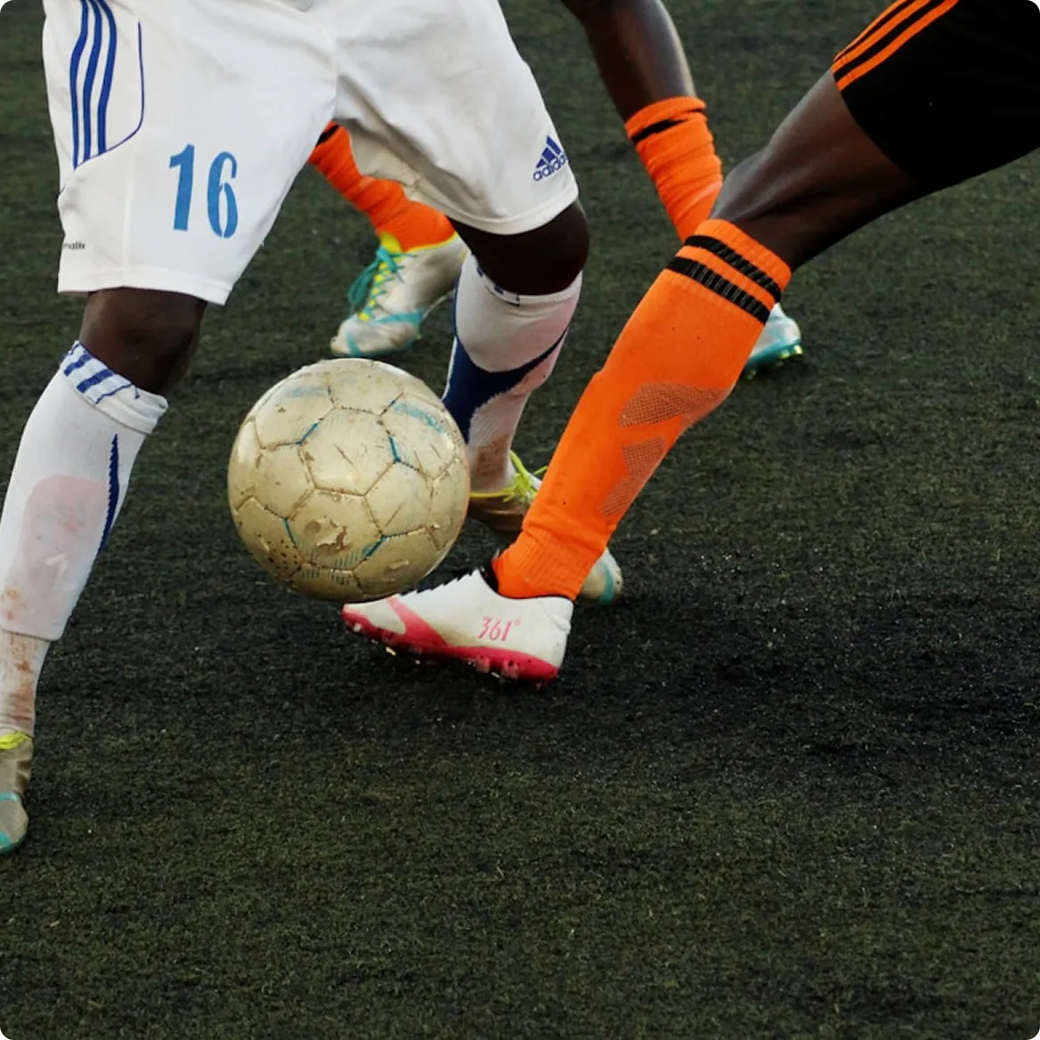 Two soccer players contesting for a worn ball on a black turf, one in white and one in orange socks.
