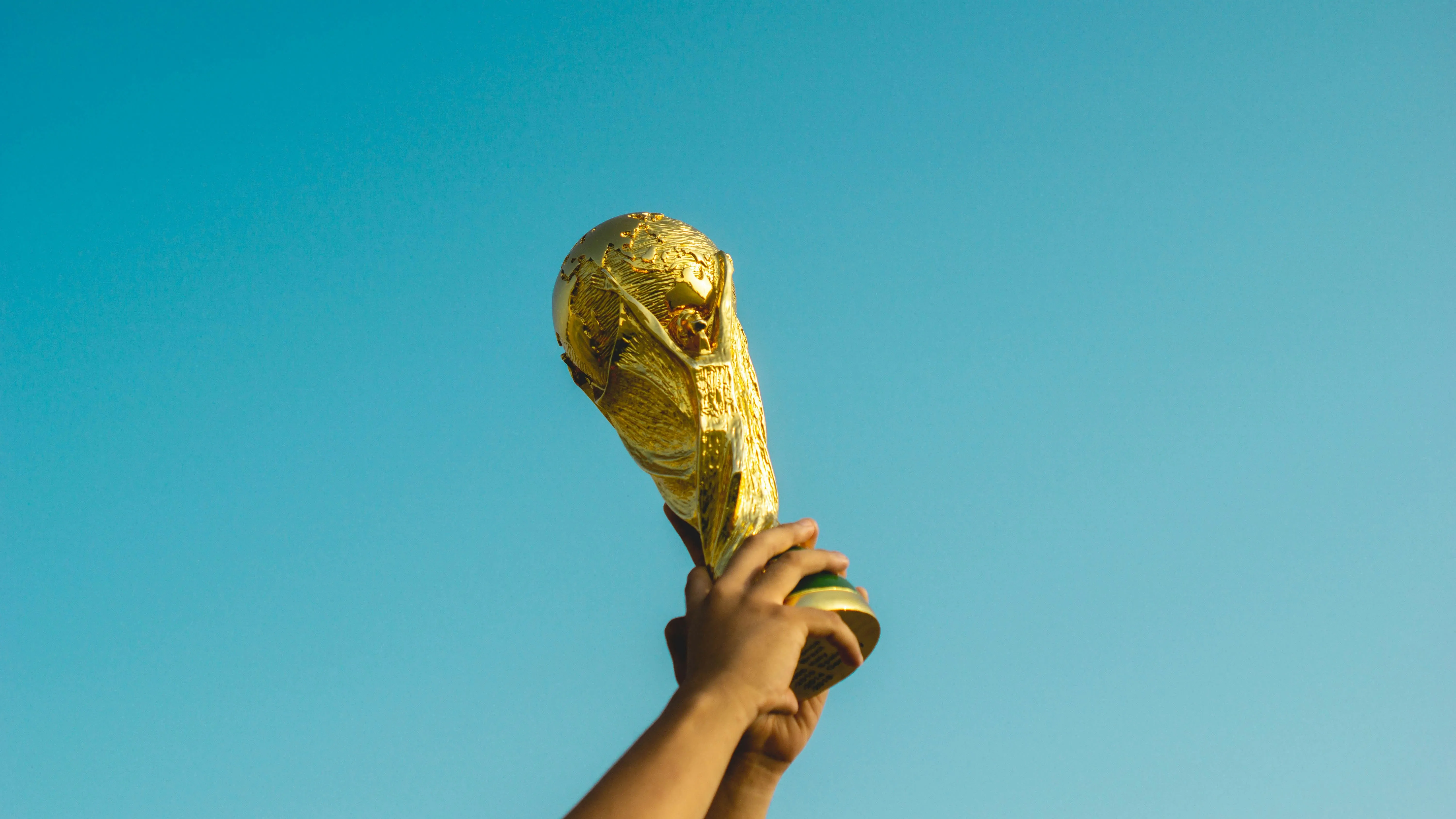Hands holding up a golden World Cup trophy against a clear blue sky.