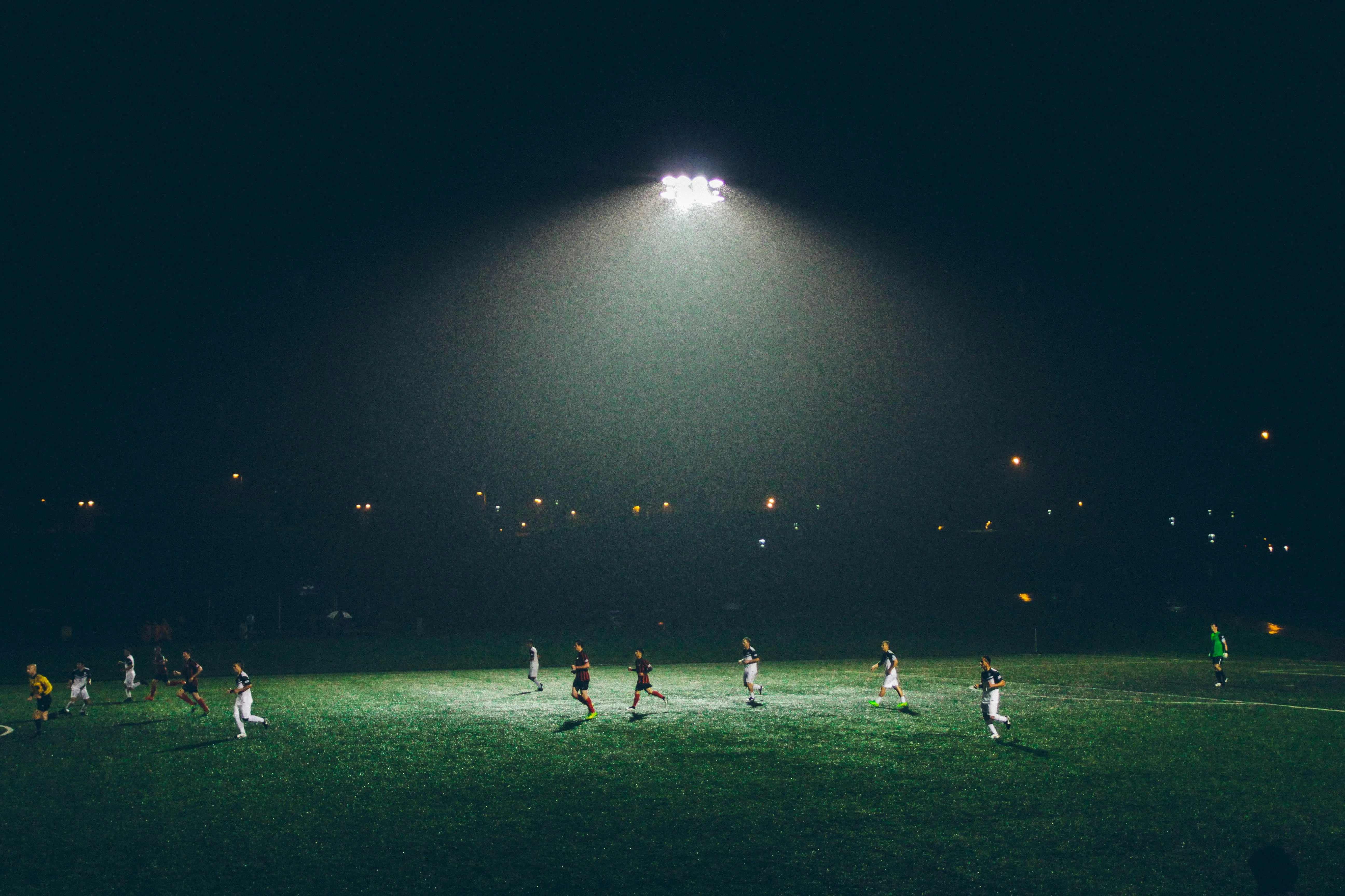 Night soccer game on wet field under bright stadium lights with players in dark and white jerseys.