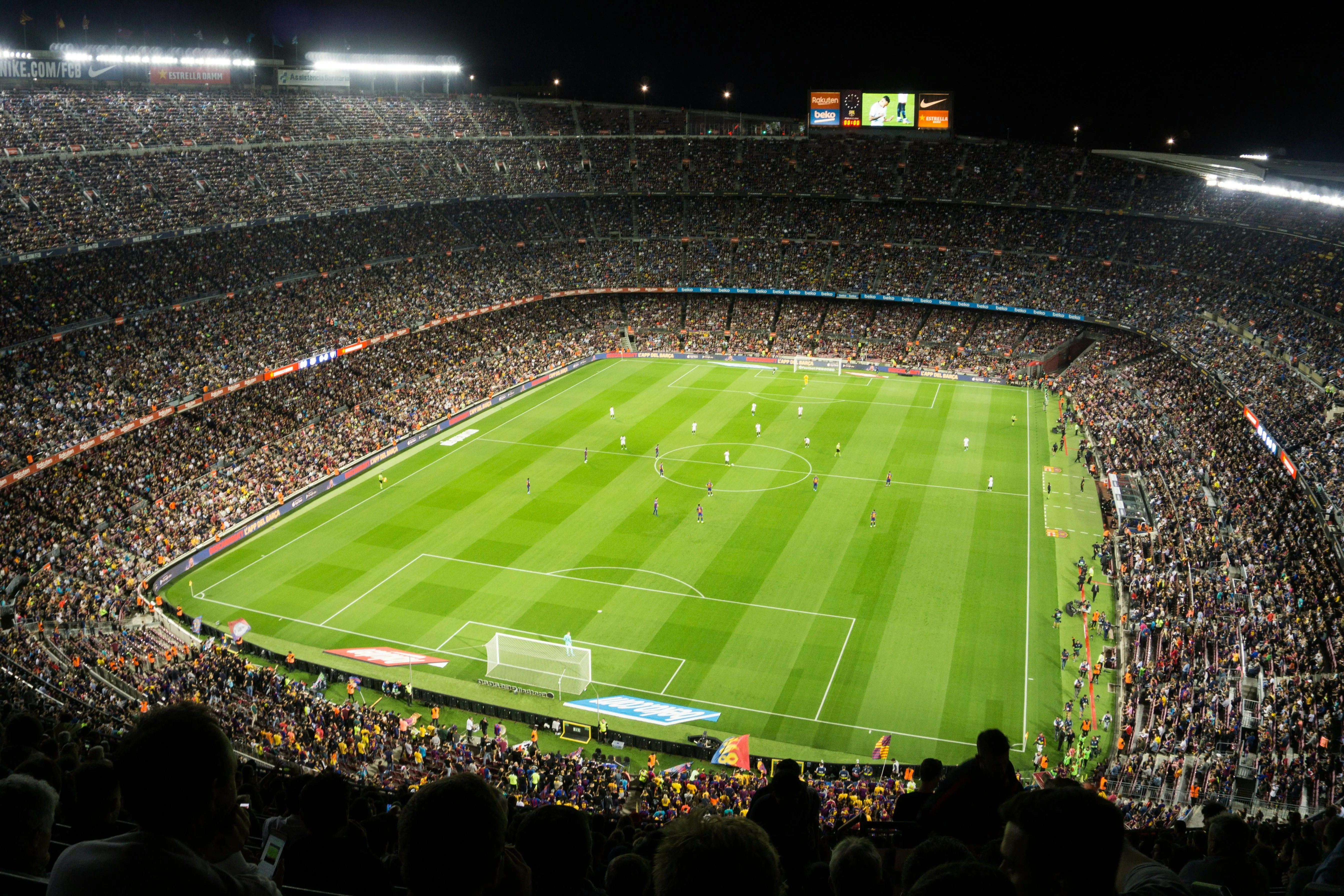 Nighttime soccer match at a packed stadium with players on a green field and fans in stands.