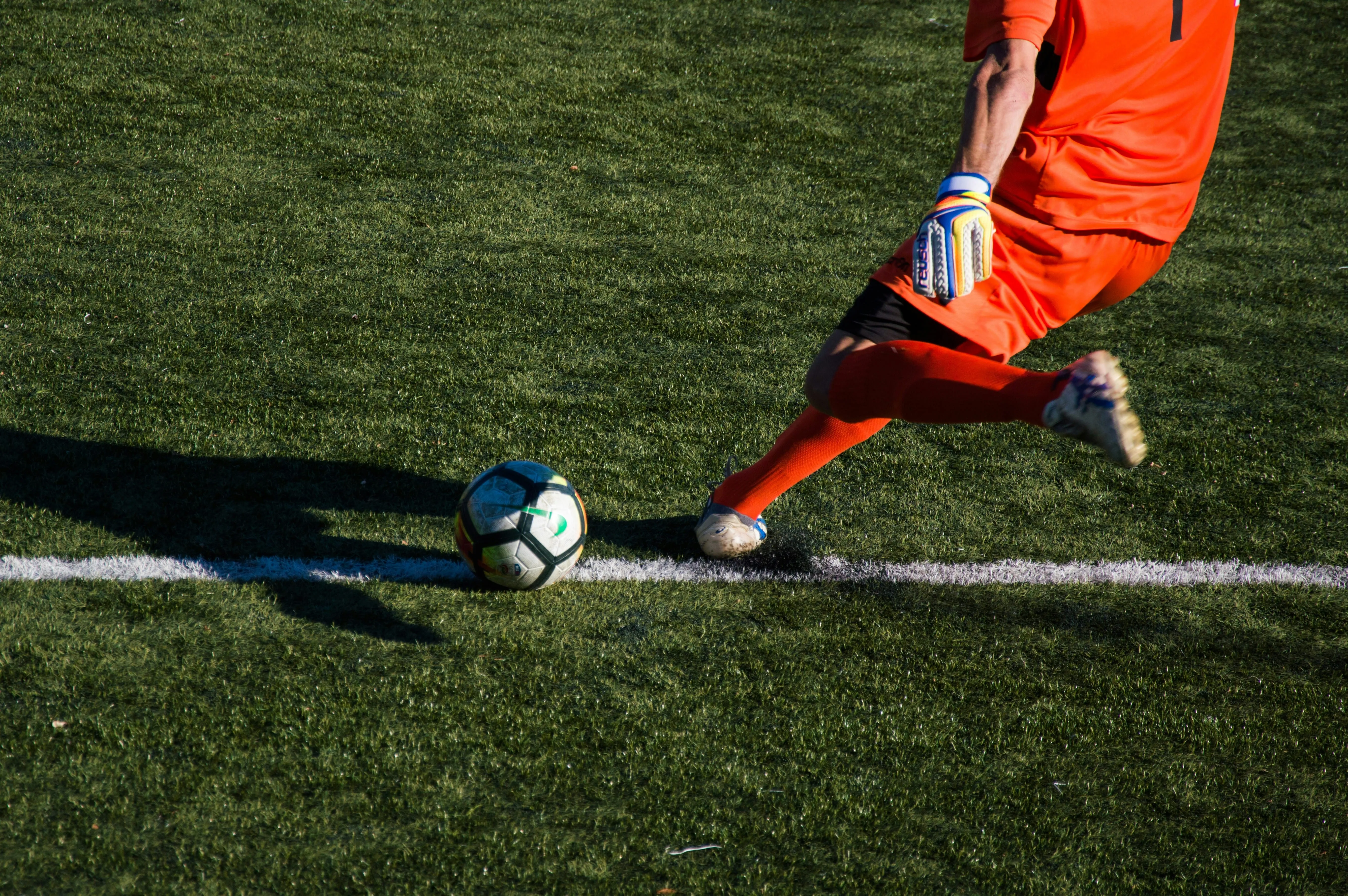 Soccer goalkeeper in orange kicking a soccer ball near the white boundary line on grass field.