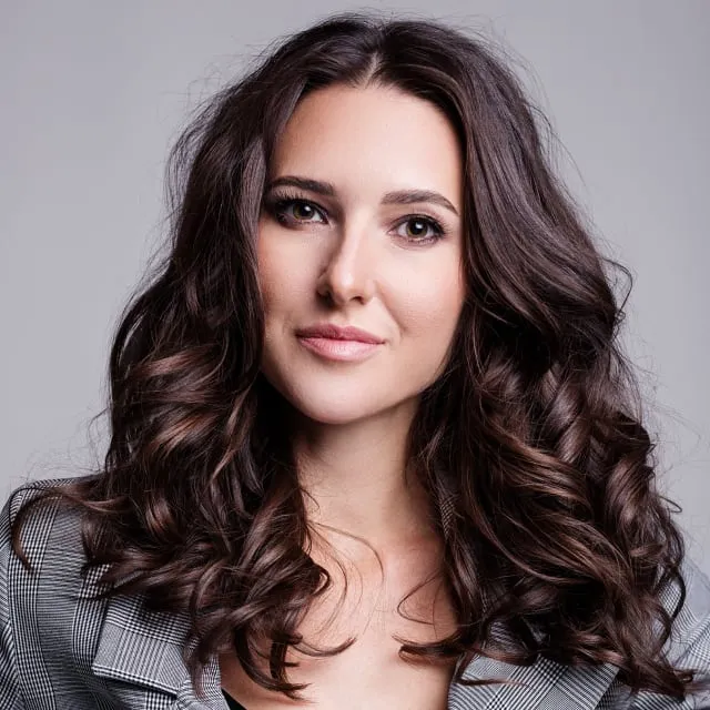 Portrait of a woman with long curly brown hair wearing a gray patterned blazer, smiling softly.