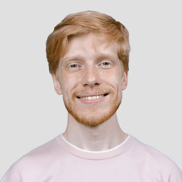 Smiling red-haired man with light beard wearing a pink shirt against plain light background.