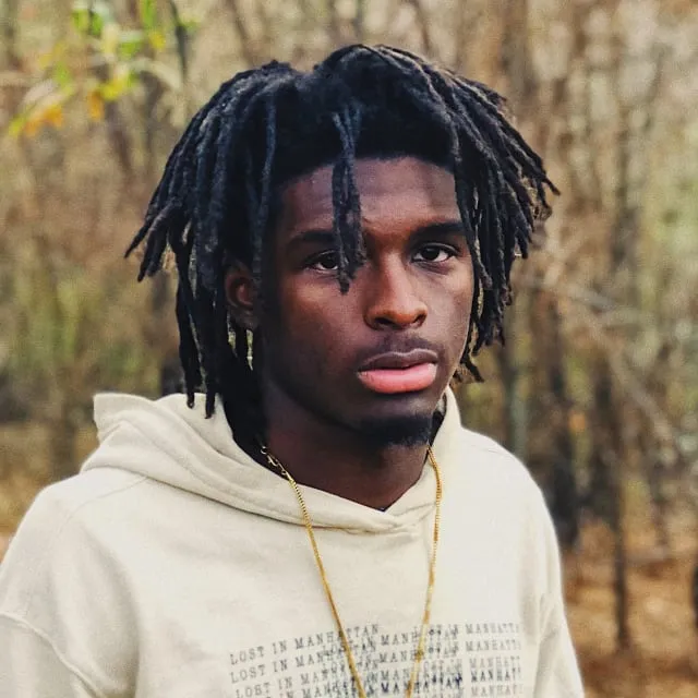 Young man with dreadlocks wearing a beige hoodie and gold chain outdoors with blurred trees behind him.