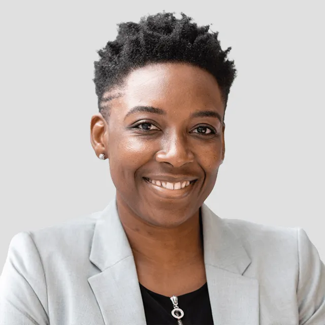 Smiling person with short curly hair wearing a light grey blazer and black top against gray background.
