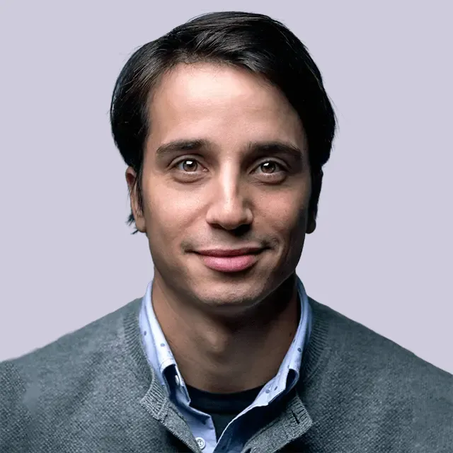 Portrait of a young man with dark hair wearing a grey sweater and collared shirt against light background.