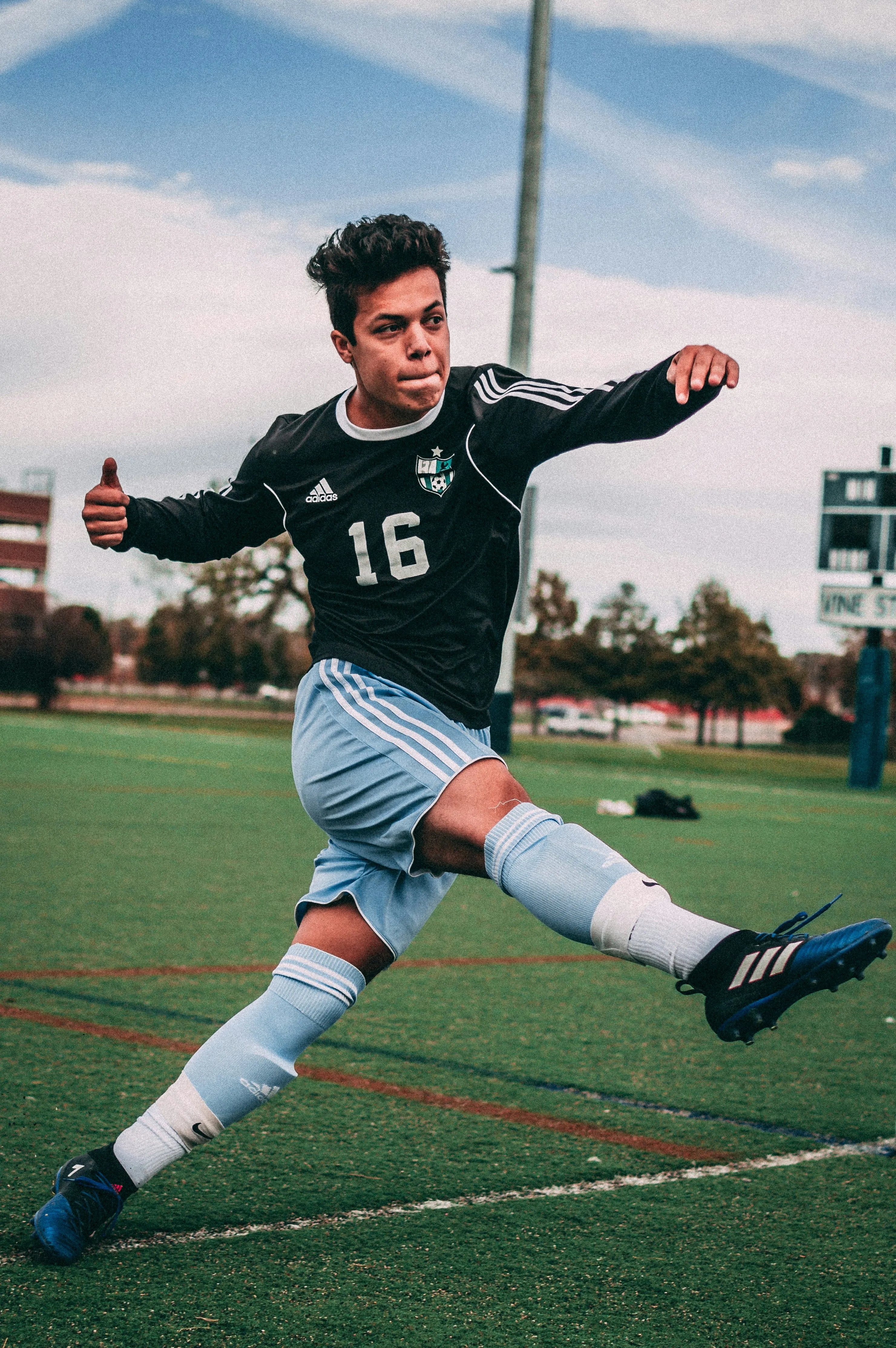 Soccer player in black and light blue kit kicking ball on green field during daytime.