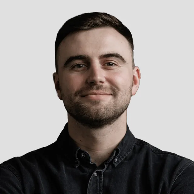 Smiling young man with short dark hair and beard wearing a black button-up shirt on gray background.