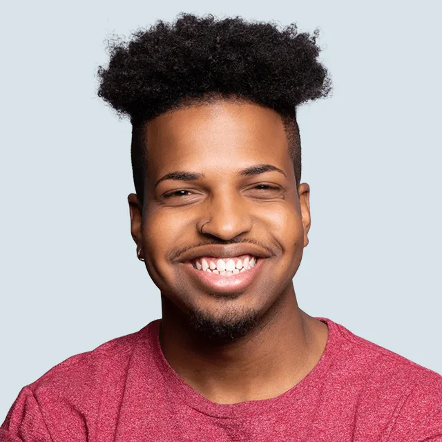 Smiling man with curly hair, nose ring, and red shirt against a light blue background.