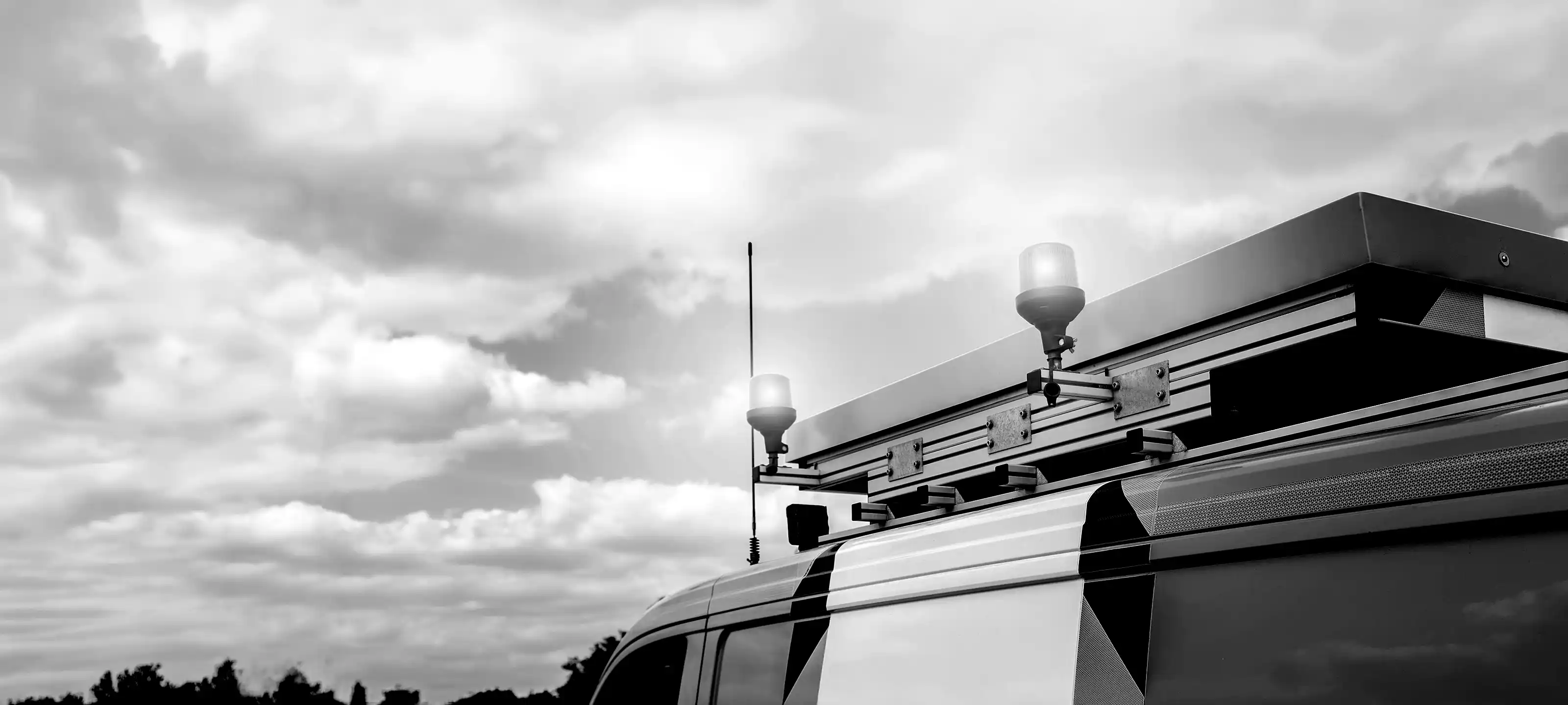 Black and white photo of the roof of an emergency vehicle with lights and an antenna under a cloudy sky.