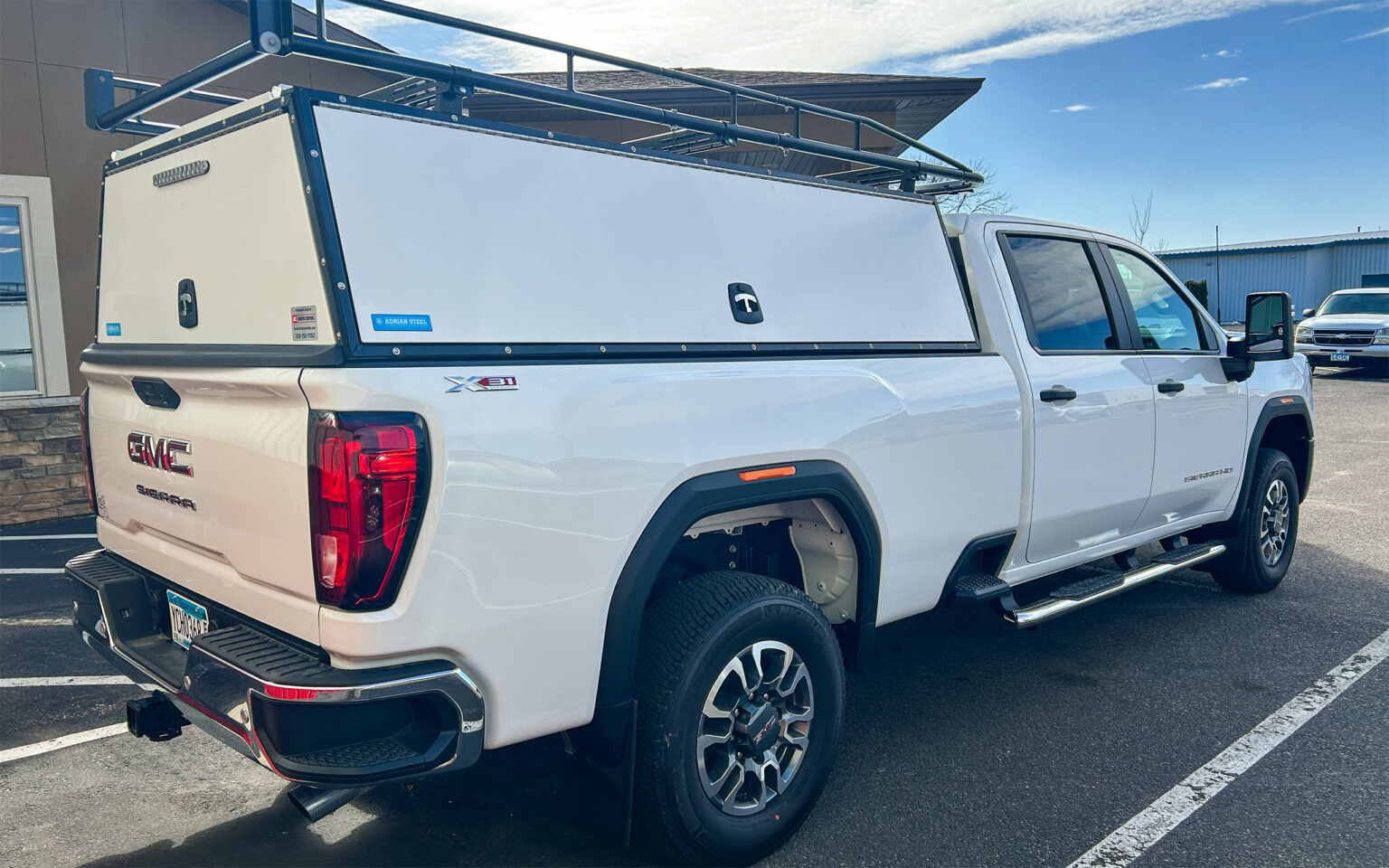 White GMC Sierra pickup truck with rooftop rack and enclosed steel bed canopy parked outdoors.