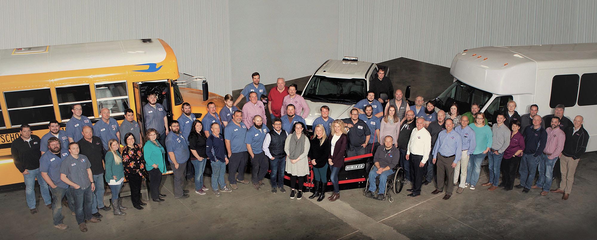 Group of people standing together in a garage with a school bus, truck, and shuttle van behind them.