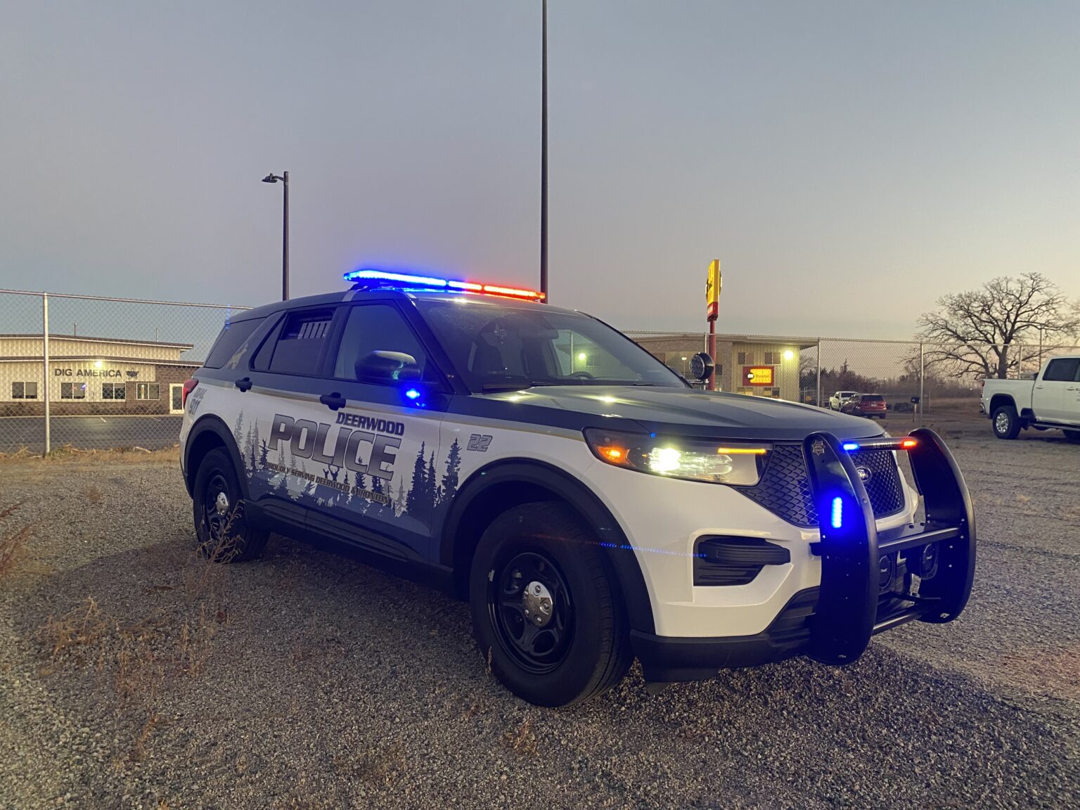 Police SUV with flashing blue and red lights parked on gravel at dusk near chain-link fence.