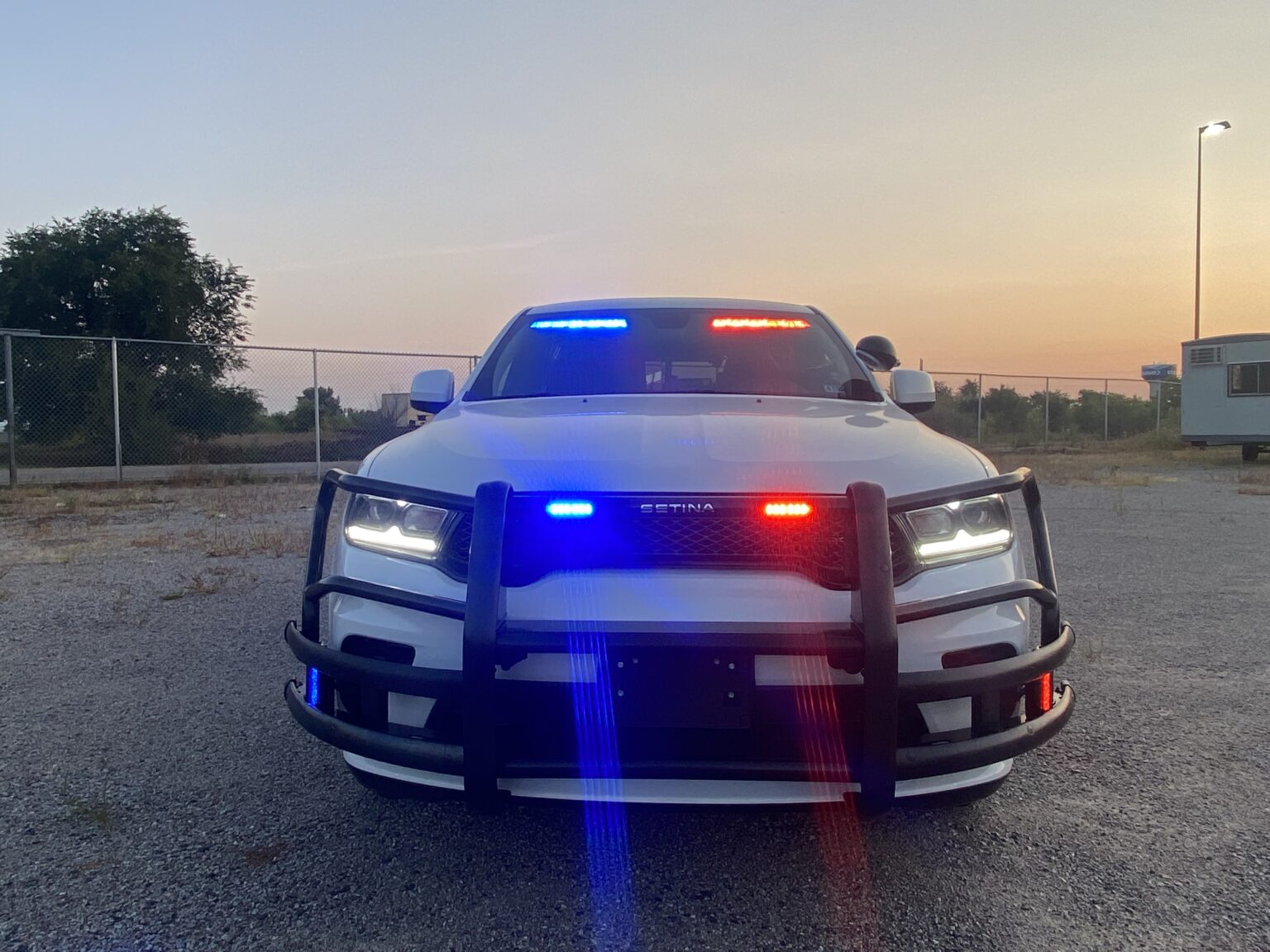 White police car with red and blue flashing lights and front protective grille at dusk.