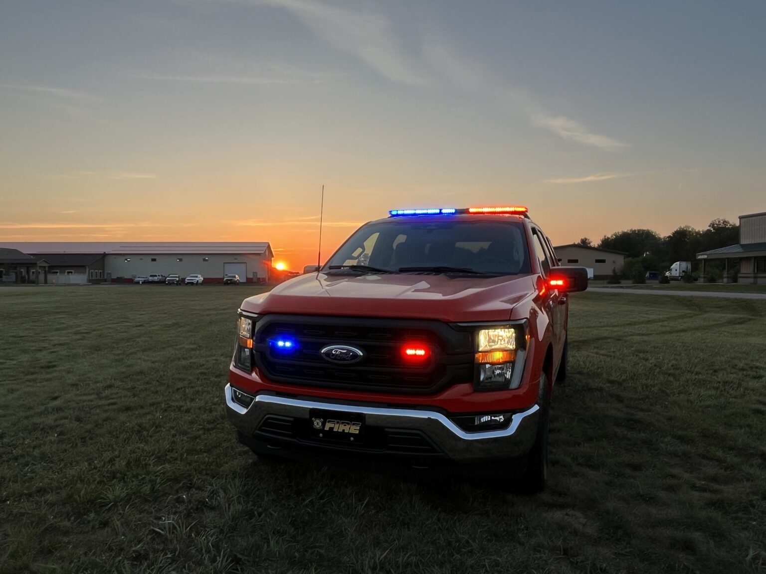 Red fire department truck with flashing red and blue lights parked on grass at sunset.