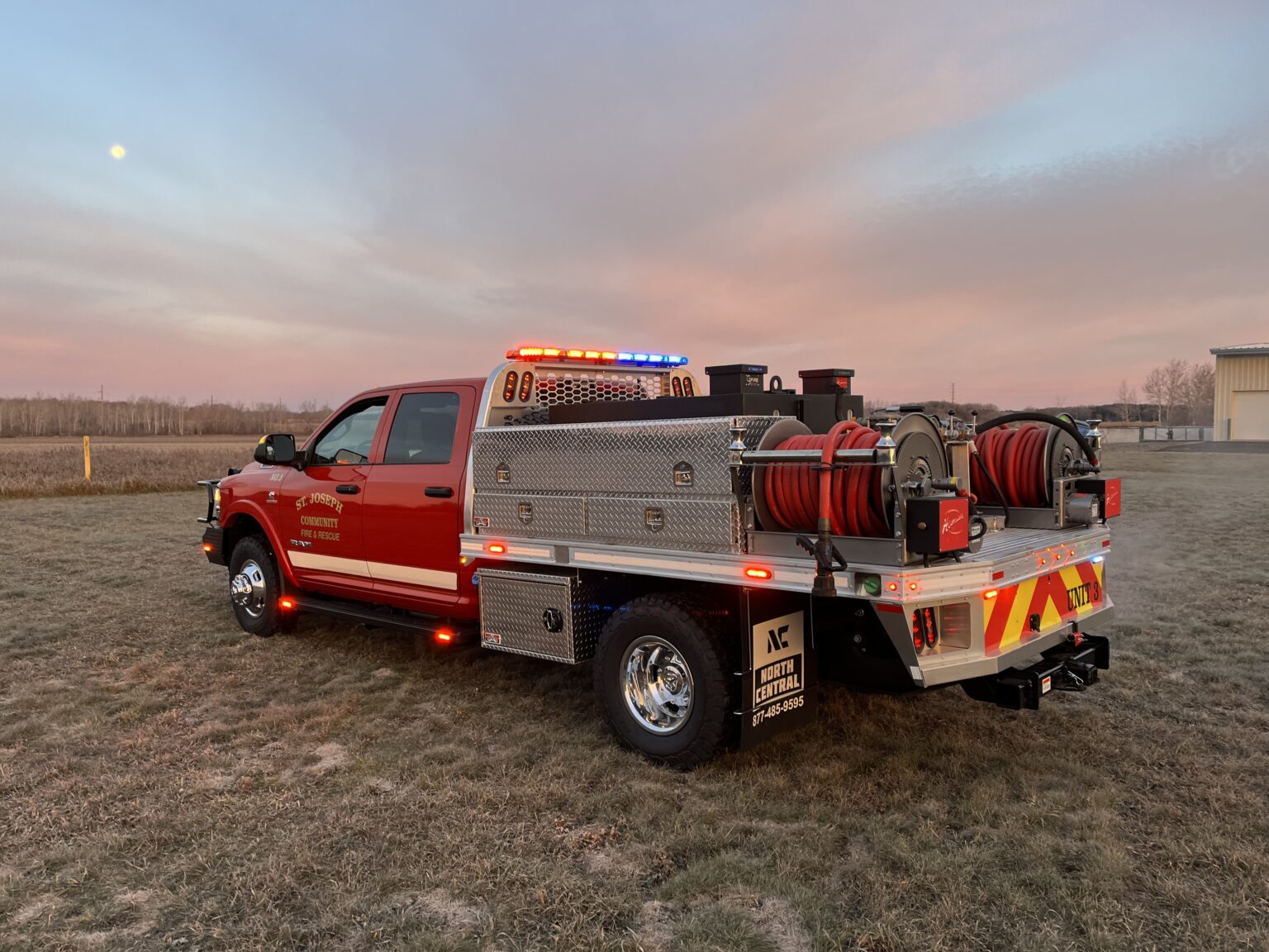 Red fire rescue truck with hoses and emergency lights parked on grass at sunset.