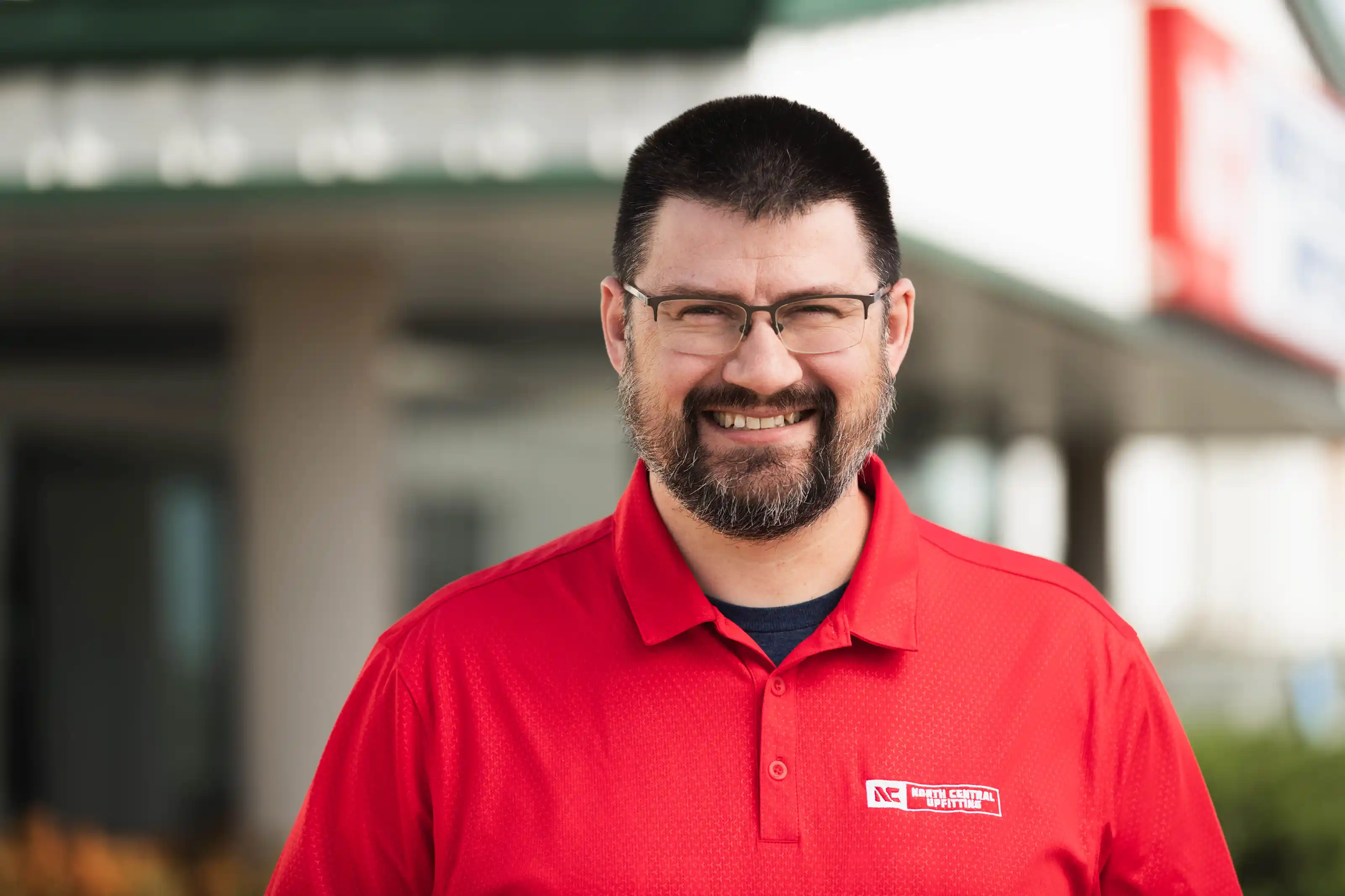 Smiling man with glasses and beard wearing a red North Central Upfitting polo shirt outdoors.