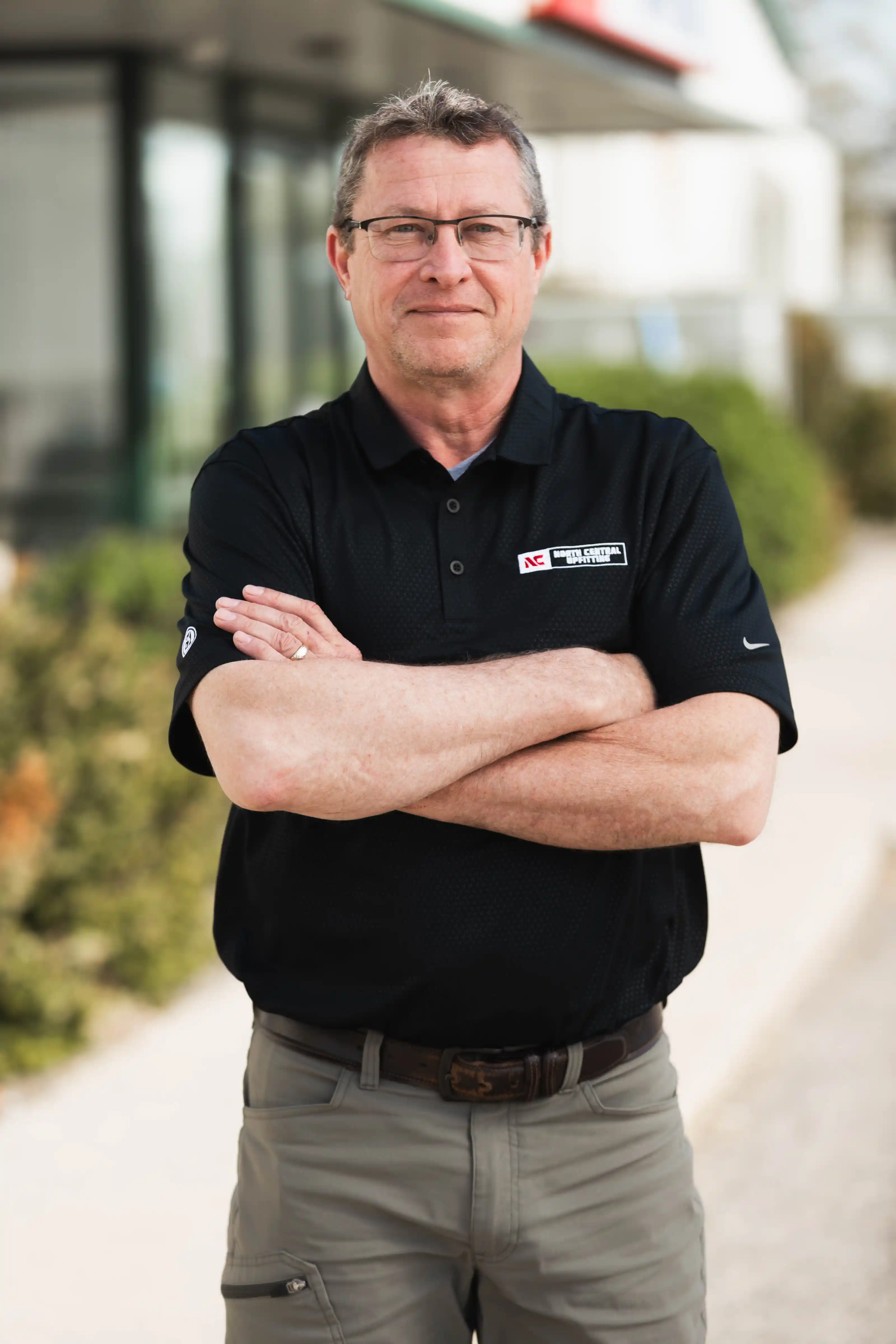 Man wearing glasses with arms crossed, dressed in black shirt and khaki pants standing outside.