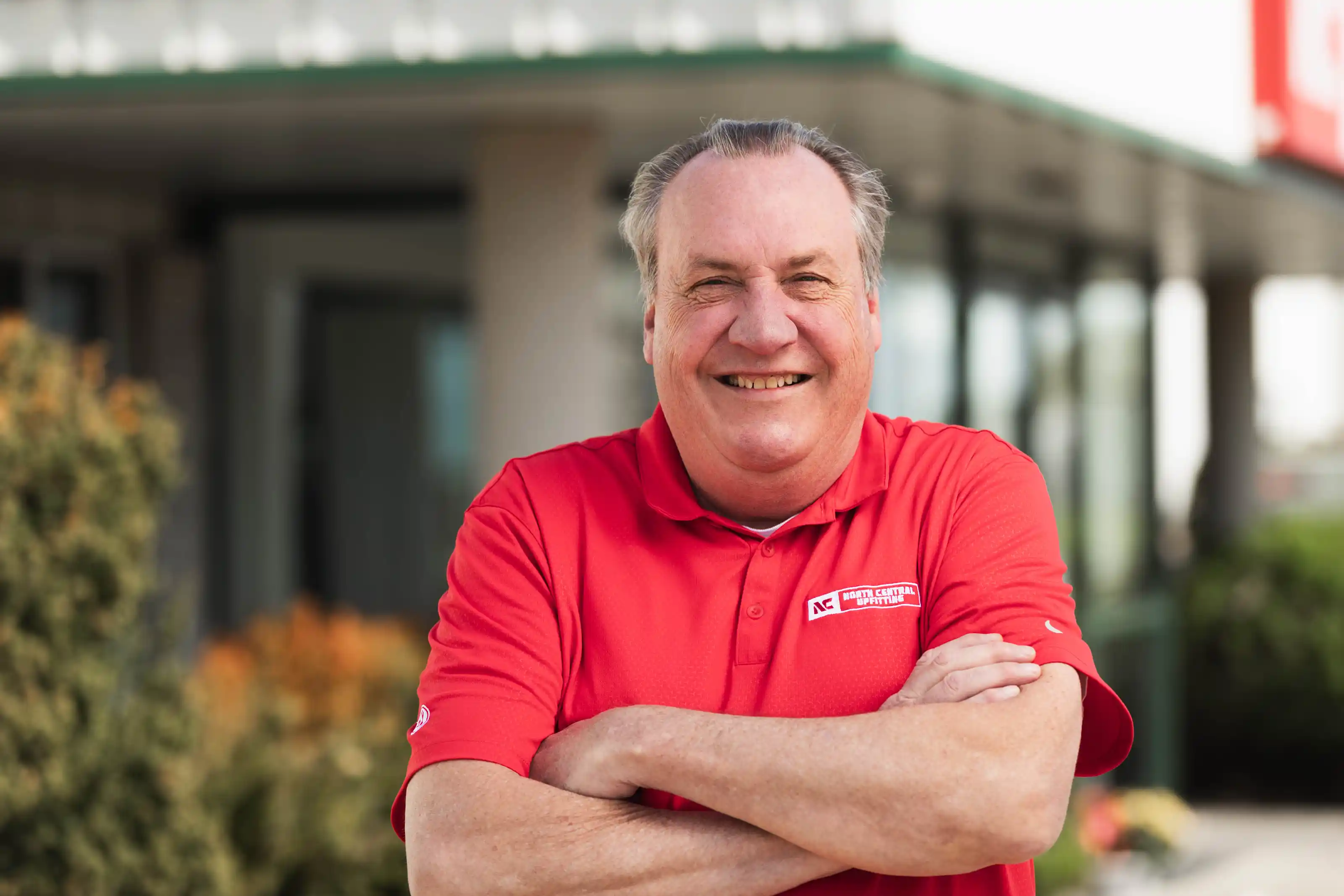 Smiling older man with folded arms wearing a red North Central Upfitting polo shirt outdoors.