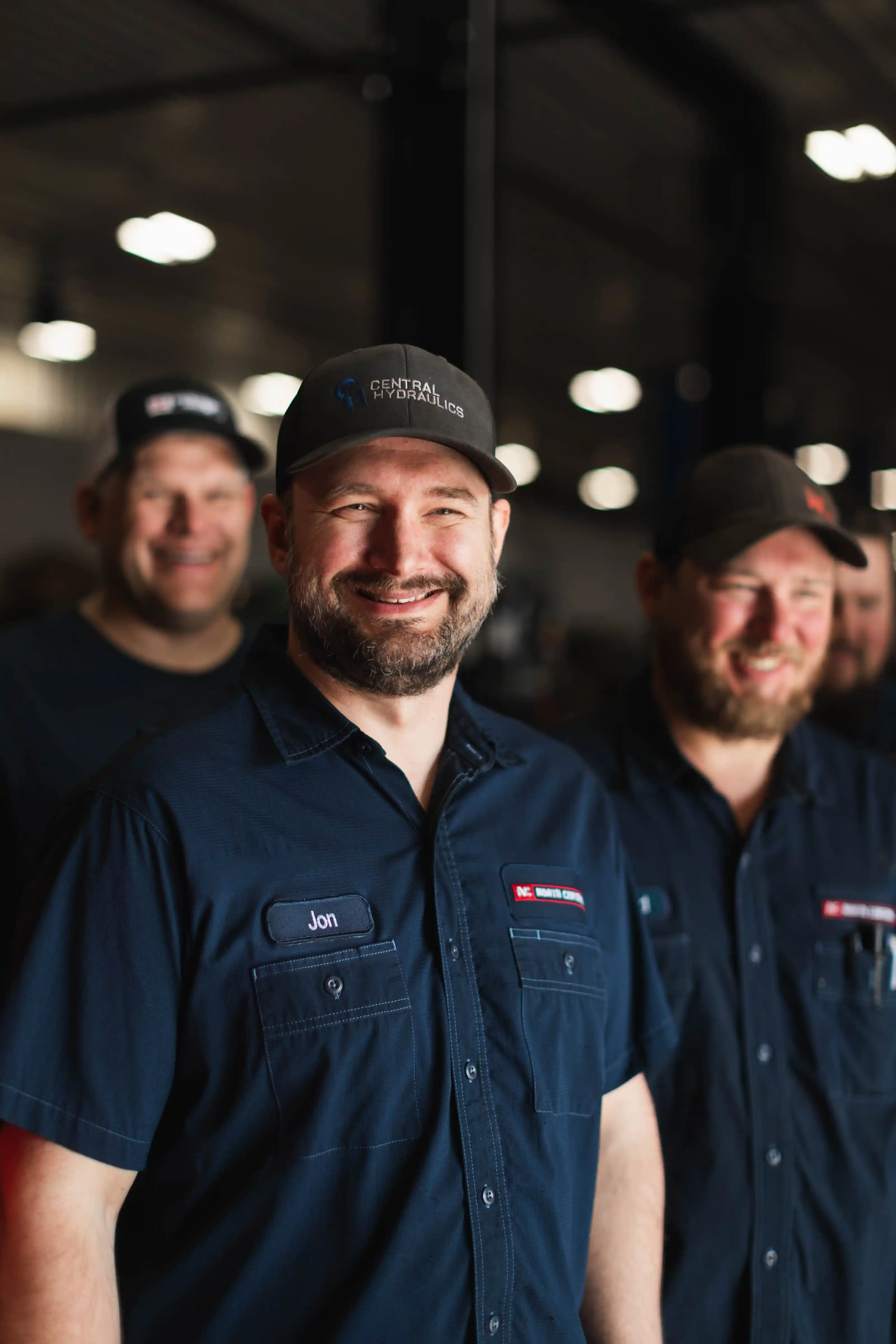 Smiling men wearing dark work shirts and caps standing in an industrial setting.