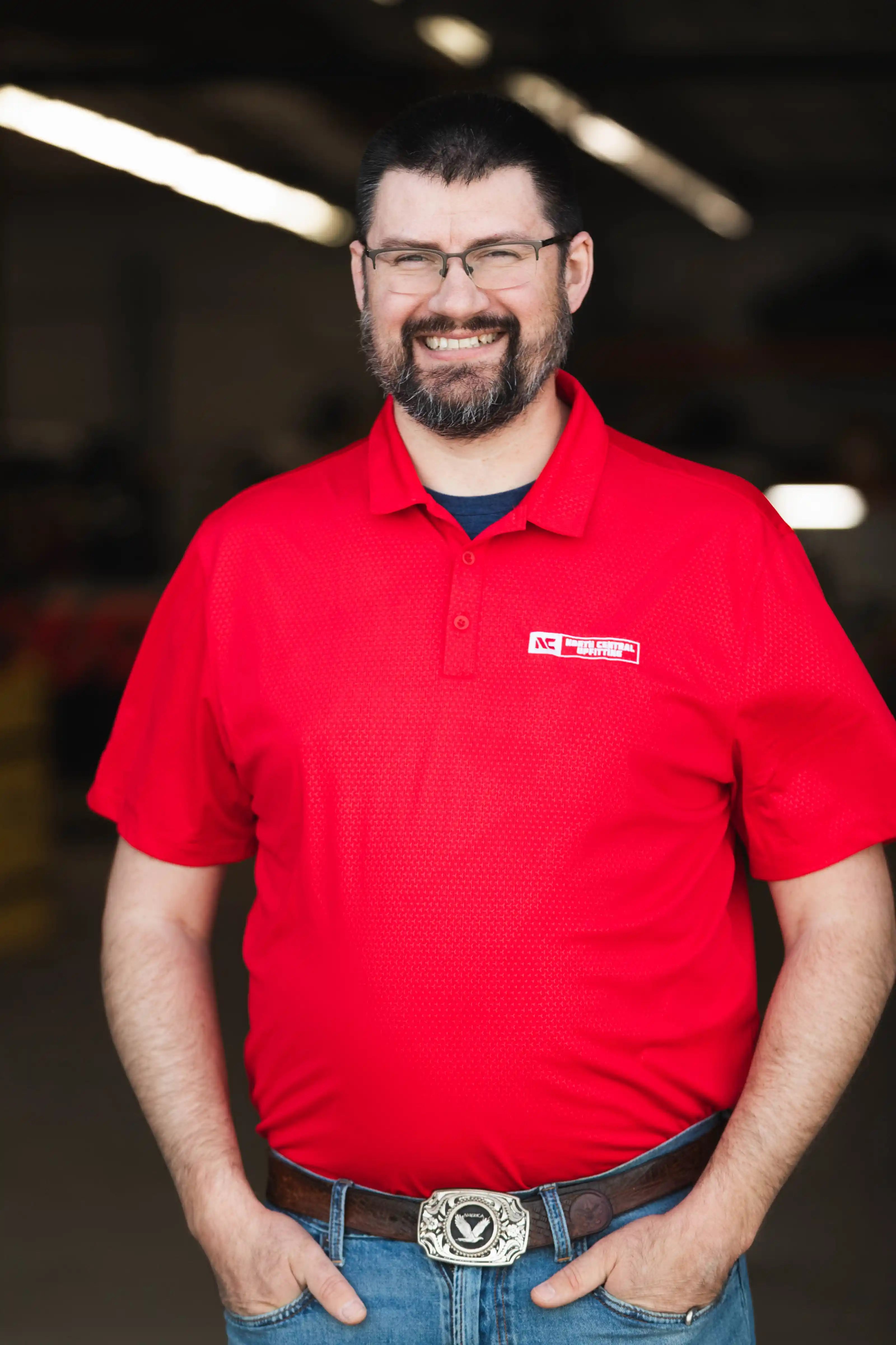 Smiling man with glasses wearing a red polo shirt and jeans, hands in pockets.