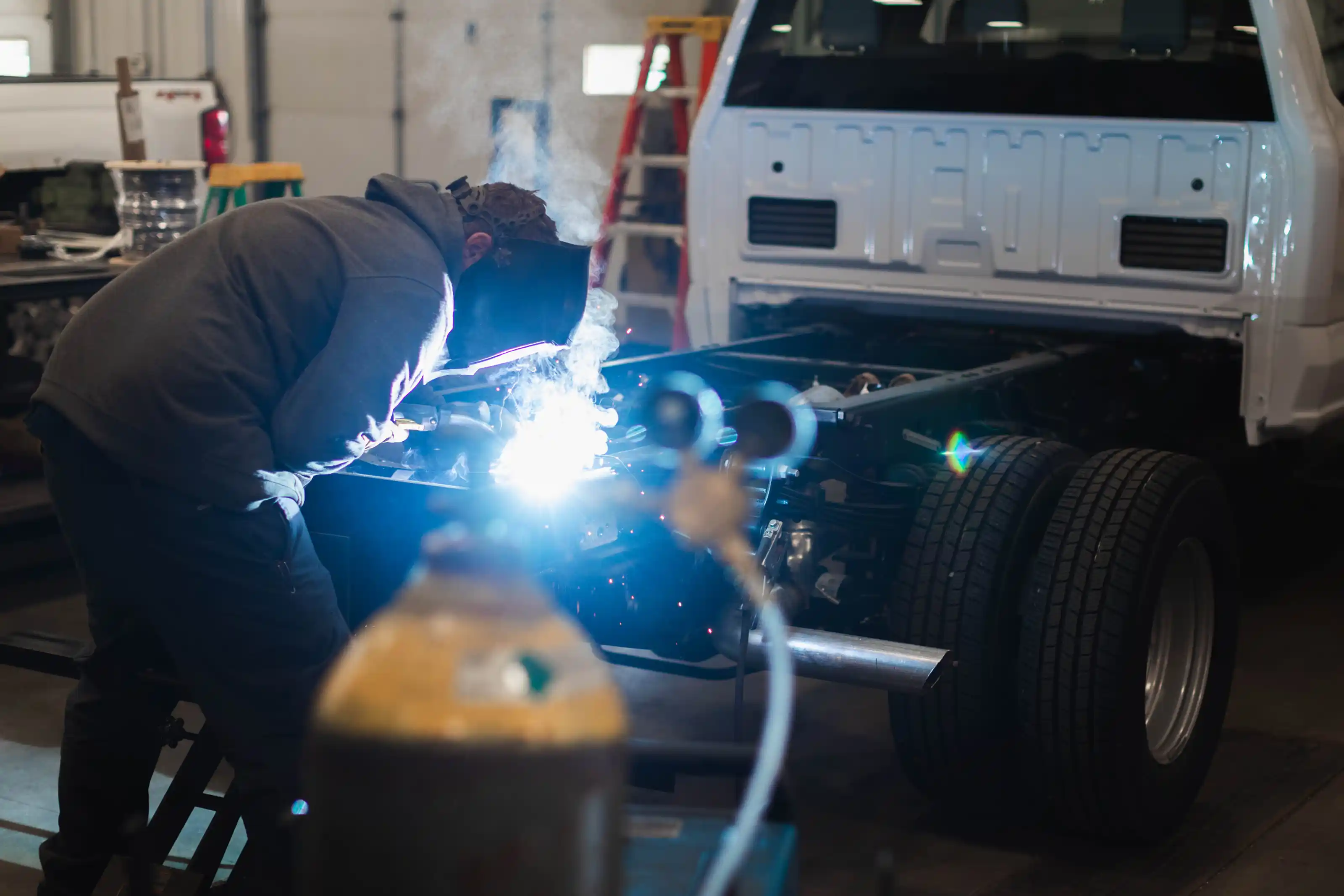 Person wearing a welding helmet and gray hoodie welding the chassis frame of a white truck inside a workshop.