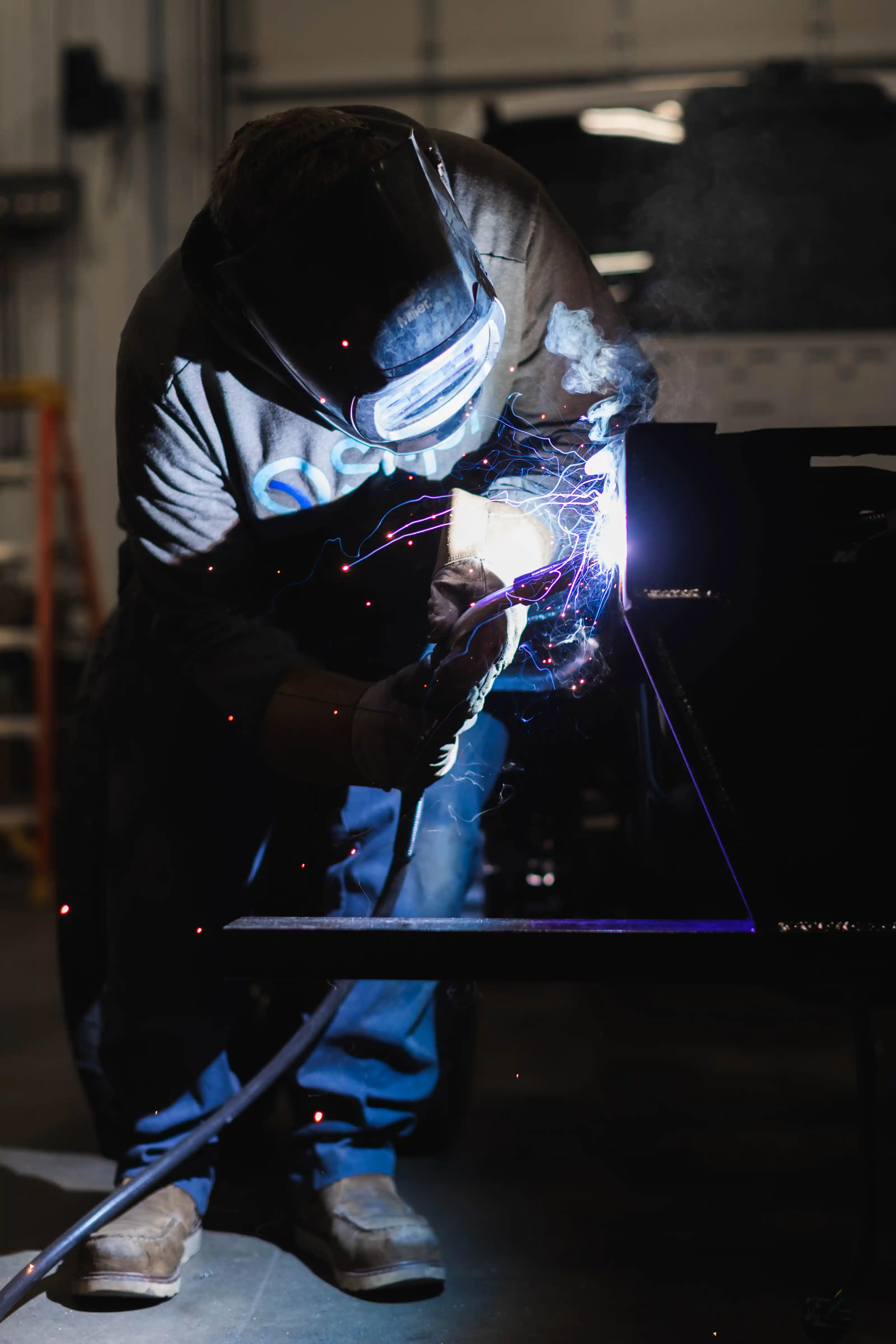 Person welding metal while wearing protective helmet and gloves in industrial workshop.