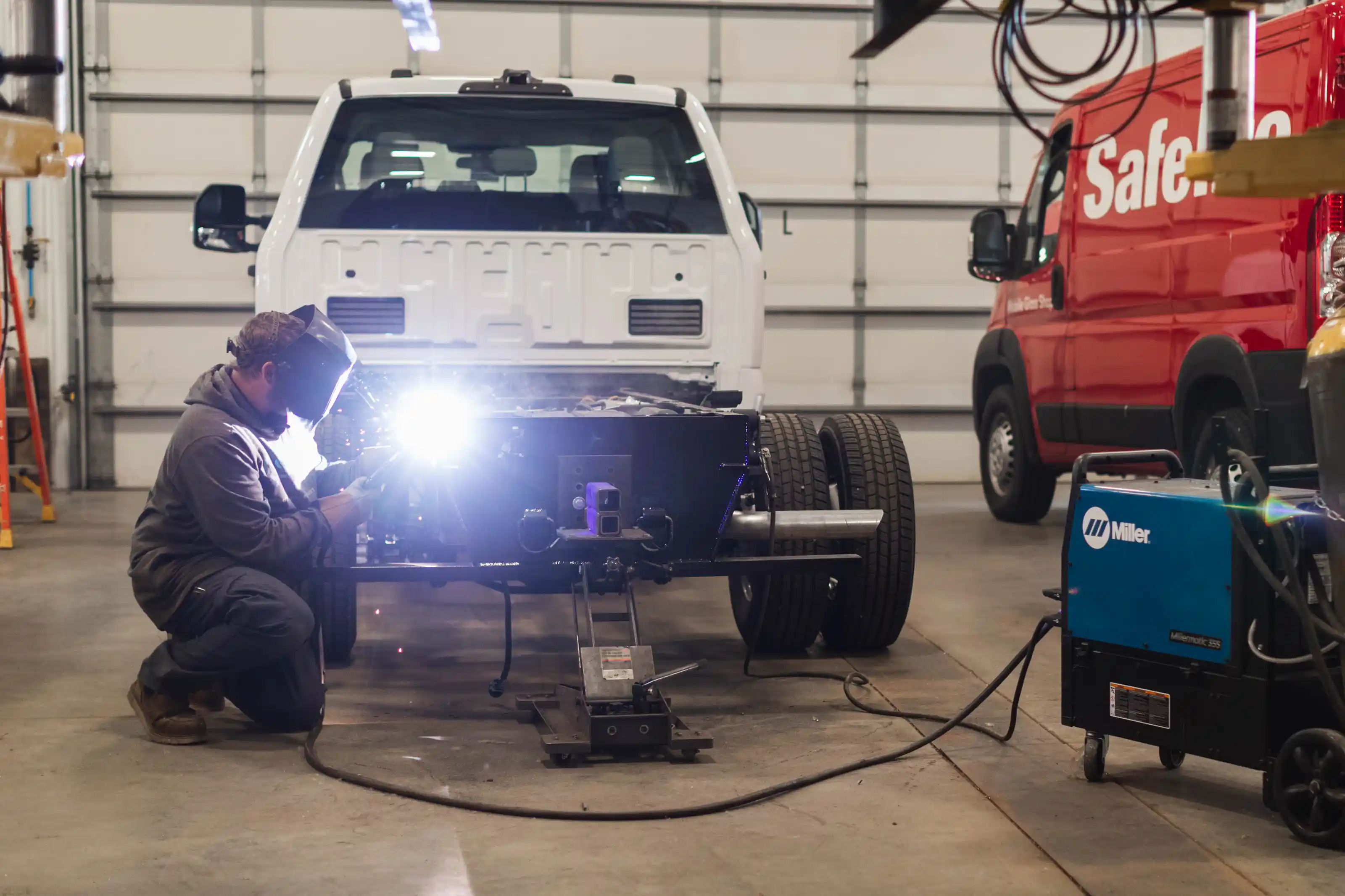 Worker welding a vehicle chassis in a garage, wearing protective gear with a Miller welding machine nearby.