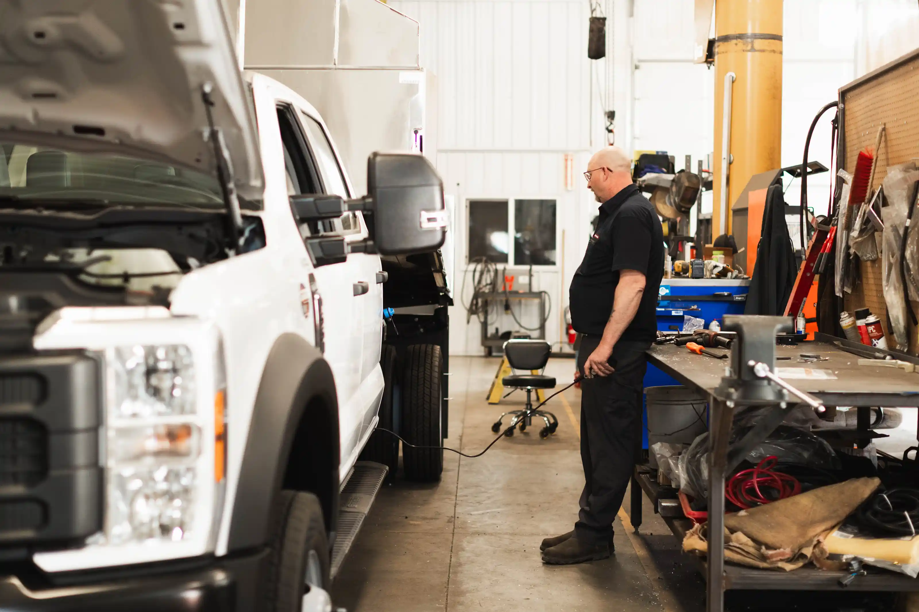 Mechanic in black uniform examining a white truck with the hood open in a workshop.