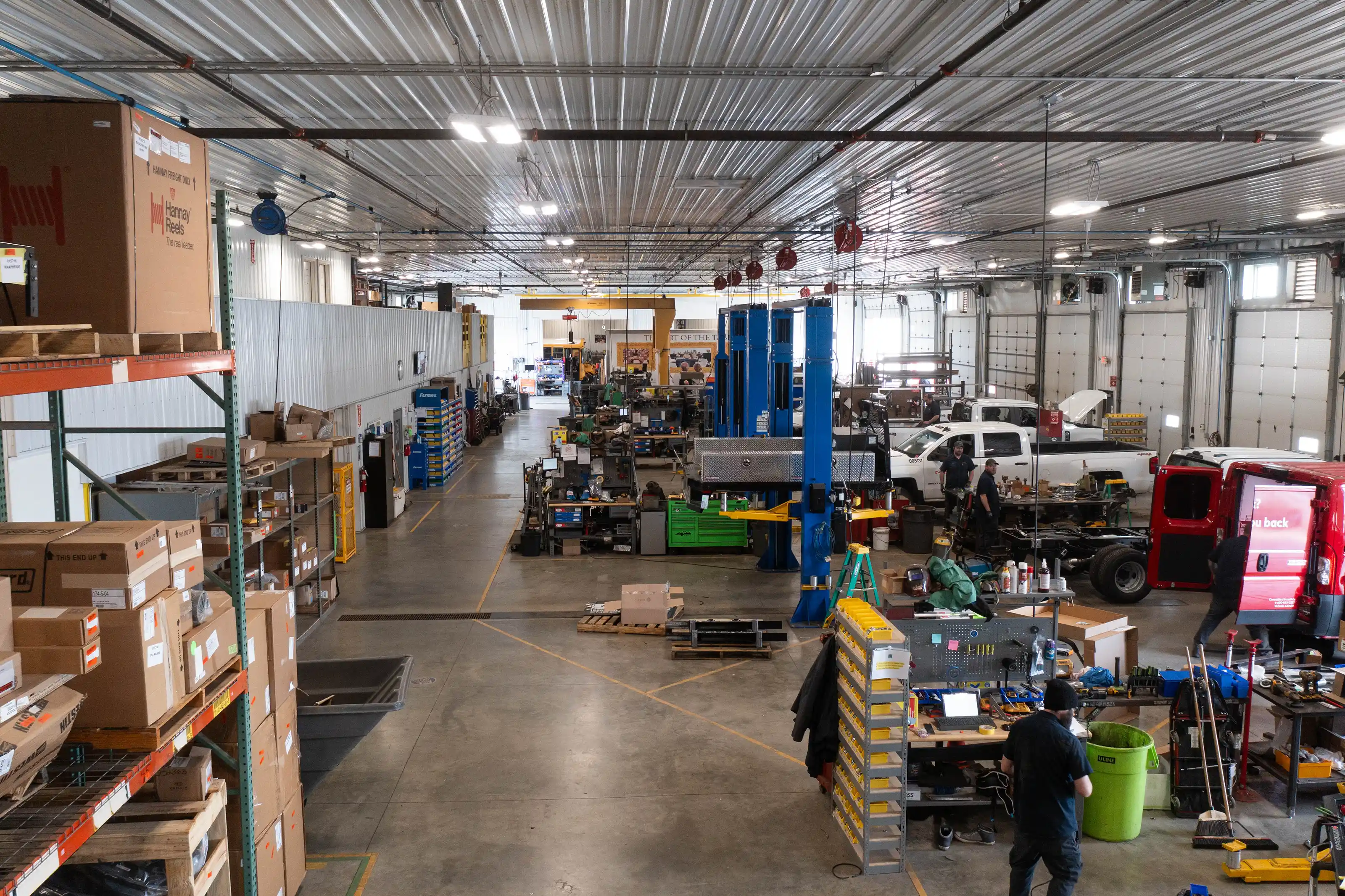 Industrial workshop with storage shelves, vehicle lifts, tools, and workers in a large garage space.