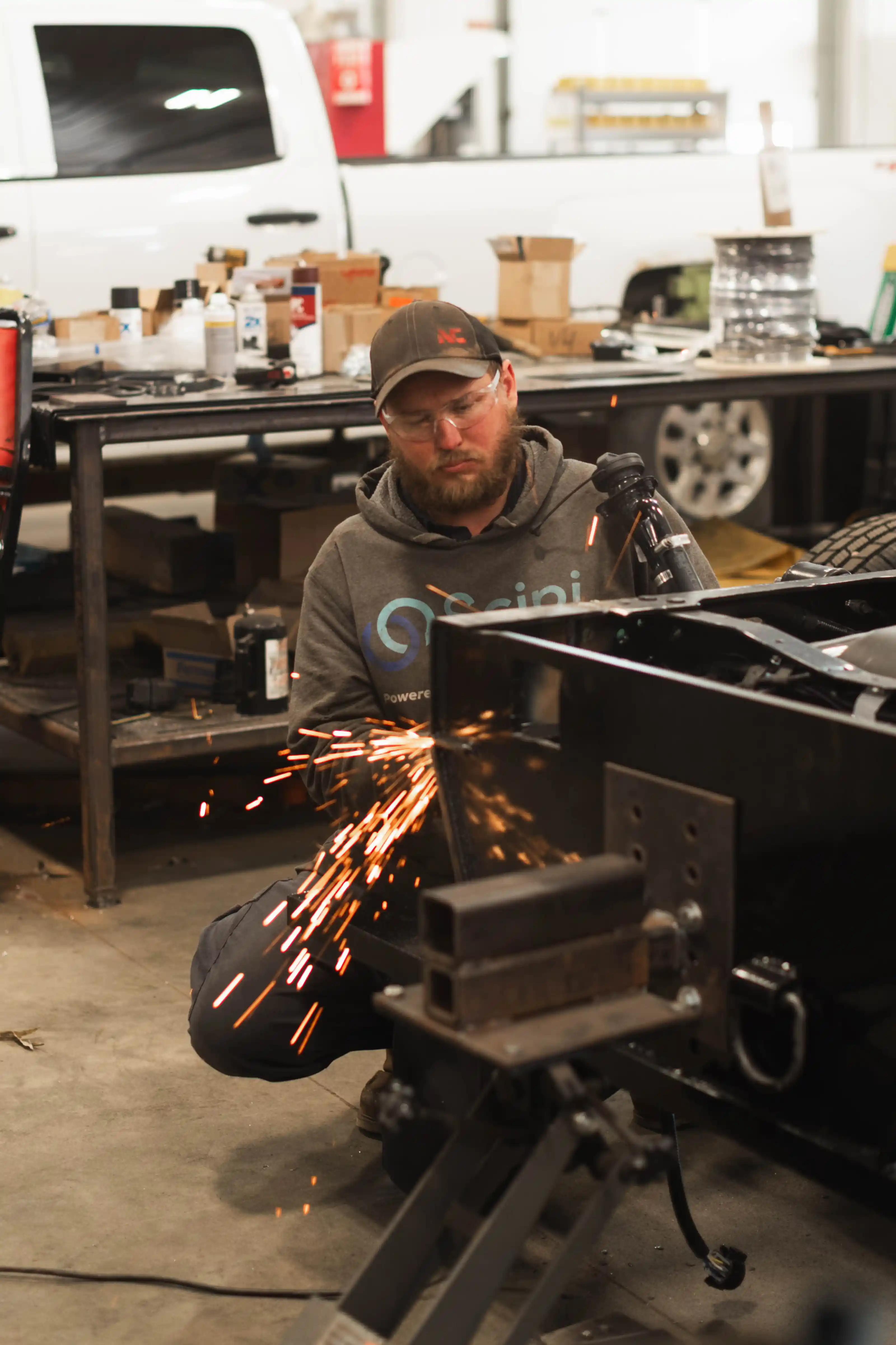 Man wearing safety glasses using a grinder producing sparks on a metal frame in a workshop.
