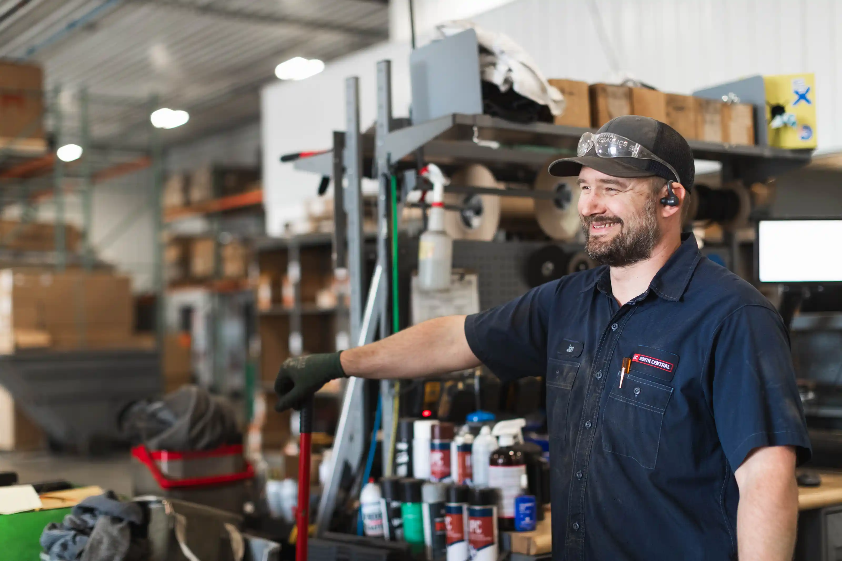 Smiling man in work uniform and cap holding a broom in an industrial warehouse setting.