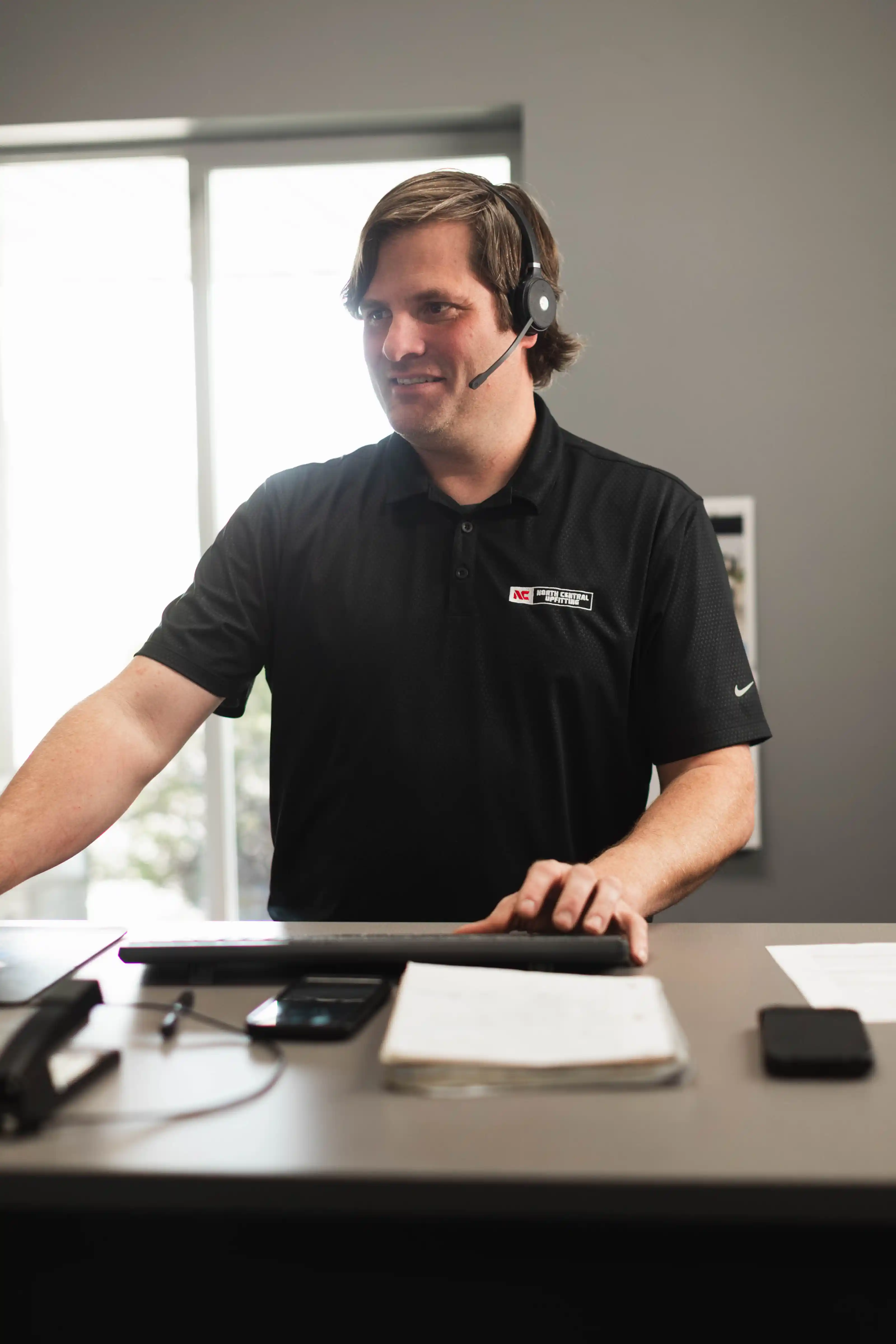 Man wearing headset and black polo shirt working at desk with keyboard, phone, and papers nearby.