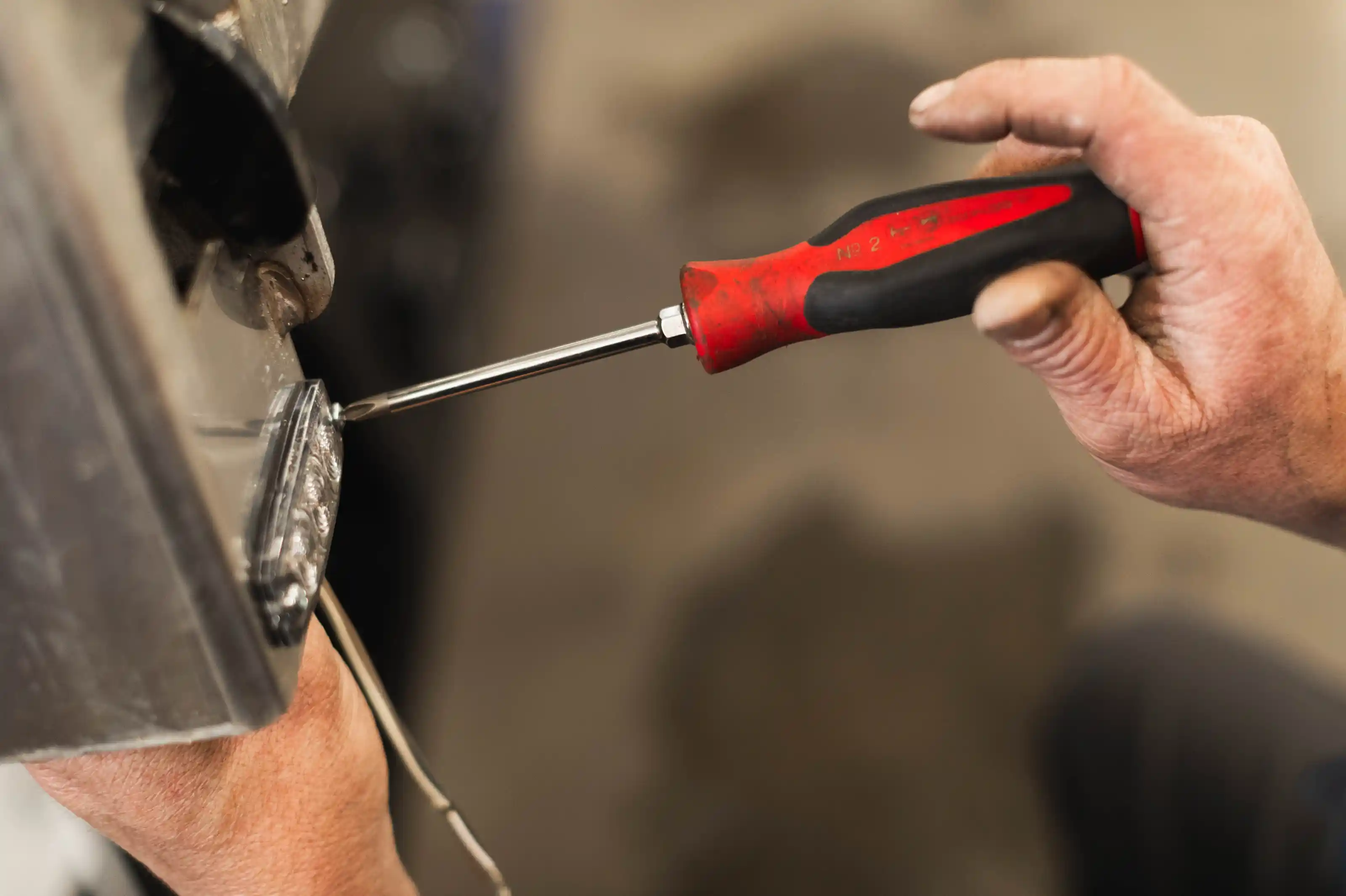 Hand using red and black screwdriver to tighten a screw on metal machinery.