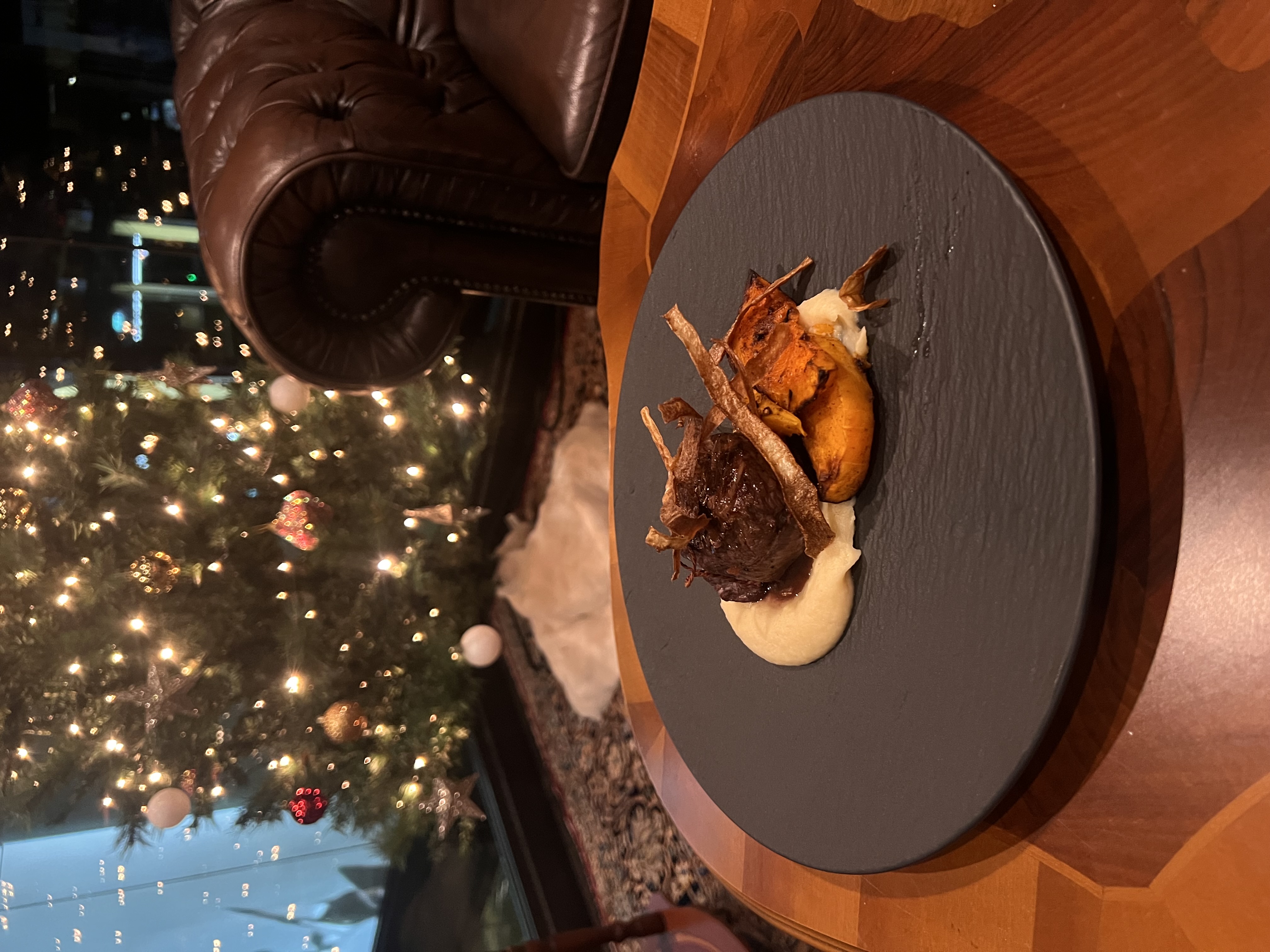 Gourmetgerecht met een stukje vlees, puree en geroosterde groenten geserveerd op een zwart bord, met een verlichte kerstboom en een leren fauteuil op de achtergrond.