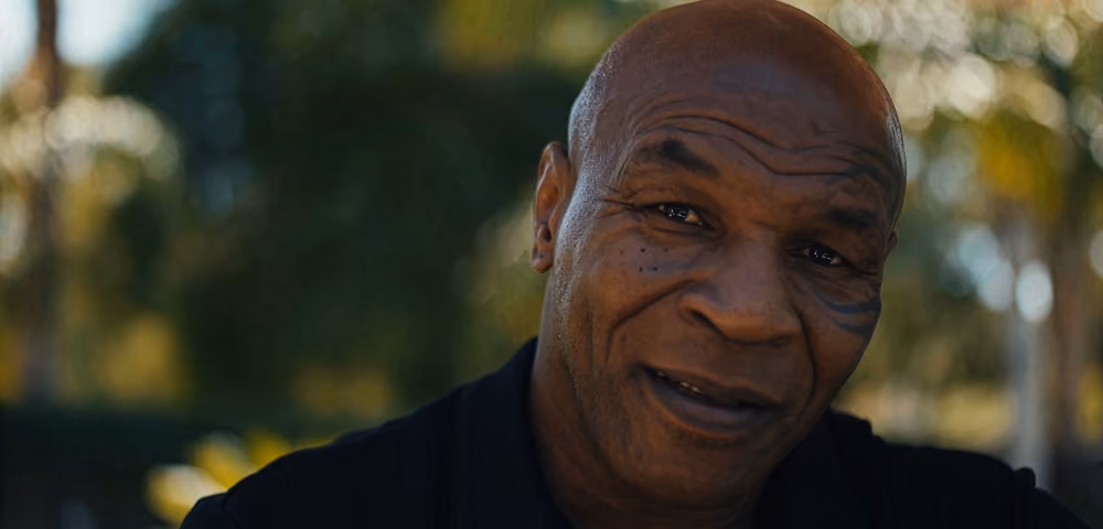 Close-up of a bald man with a facial tattoo smiling outdoors with trees in the background.