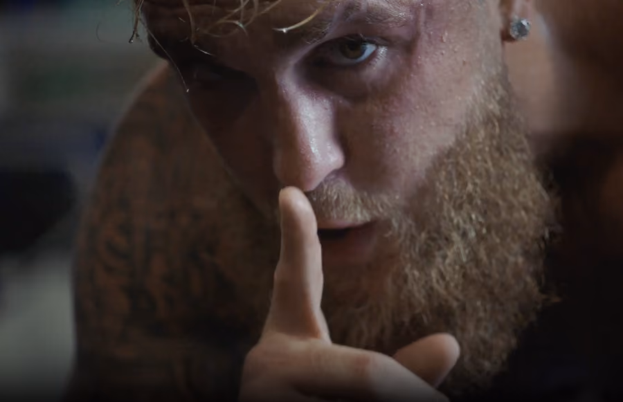 Close-up of a bearded man with a serious expression holding his finger to his lips in a shushing gesture.