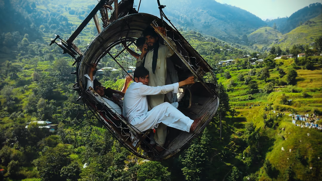Three men in traditional clothing ride a suspended basket over a green mountainous landscape.