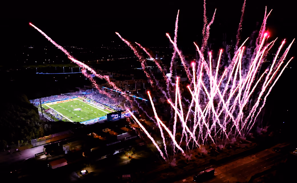Night aerial view of a stadium with fireworks lighting the sky over the field and crowd.