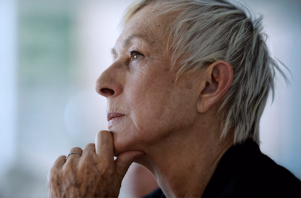 Pensive older woman with short grey hair resting chin on hand looking to the left.