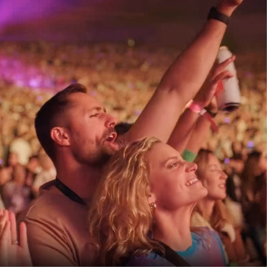 Crowd at a concert with people singing and cheering, one person holding a can raised in the air.